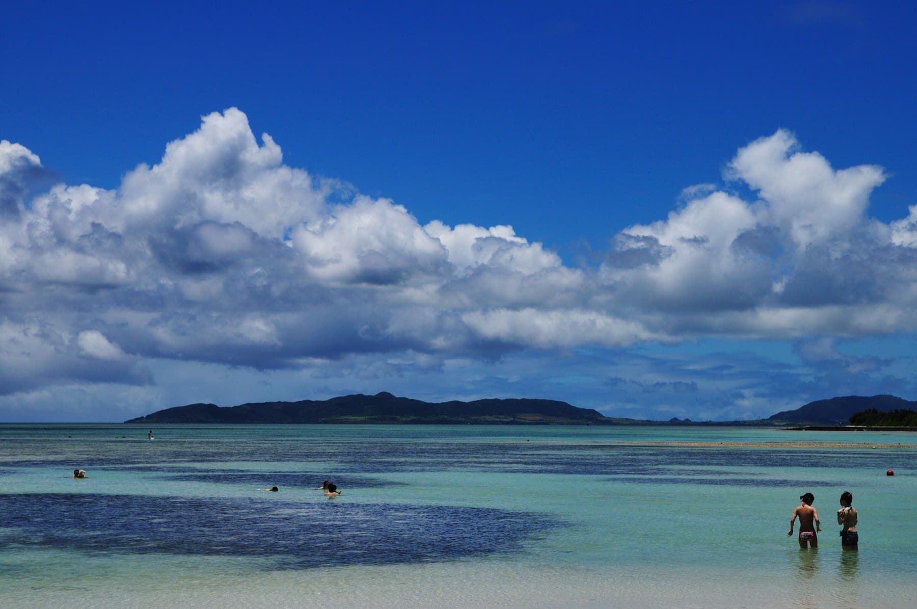 Swimmers stand waist deep in the clear shallows of Kondoi Beach as tall summer clouds build over the hills of Kohama across the channel.