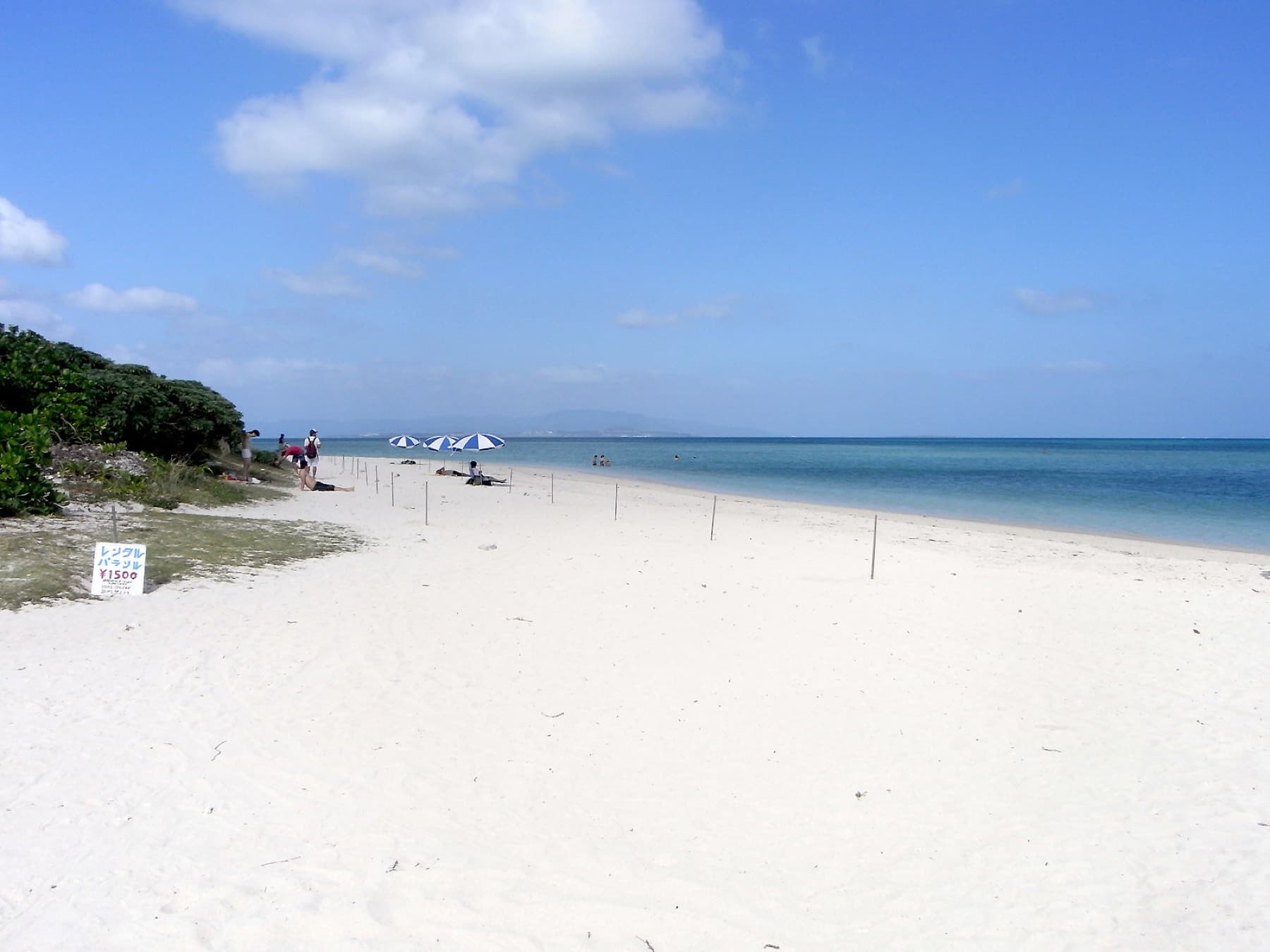 A broad sweep of white sand runs from the trees to the water at Kondoi Beach on Taketomi, where a few beach umbrellas stand near the shoreline.