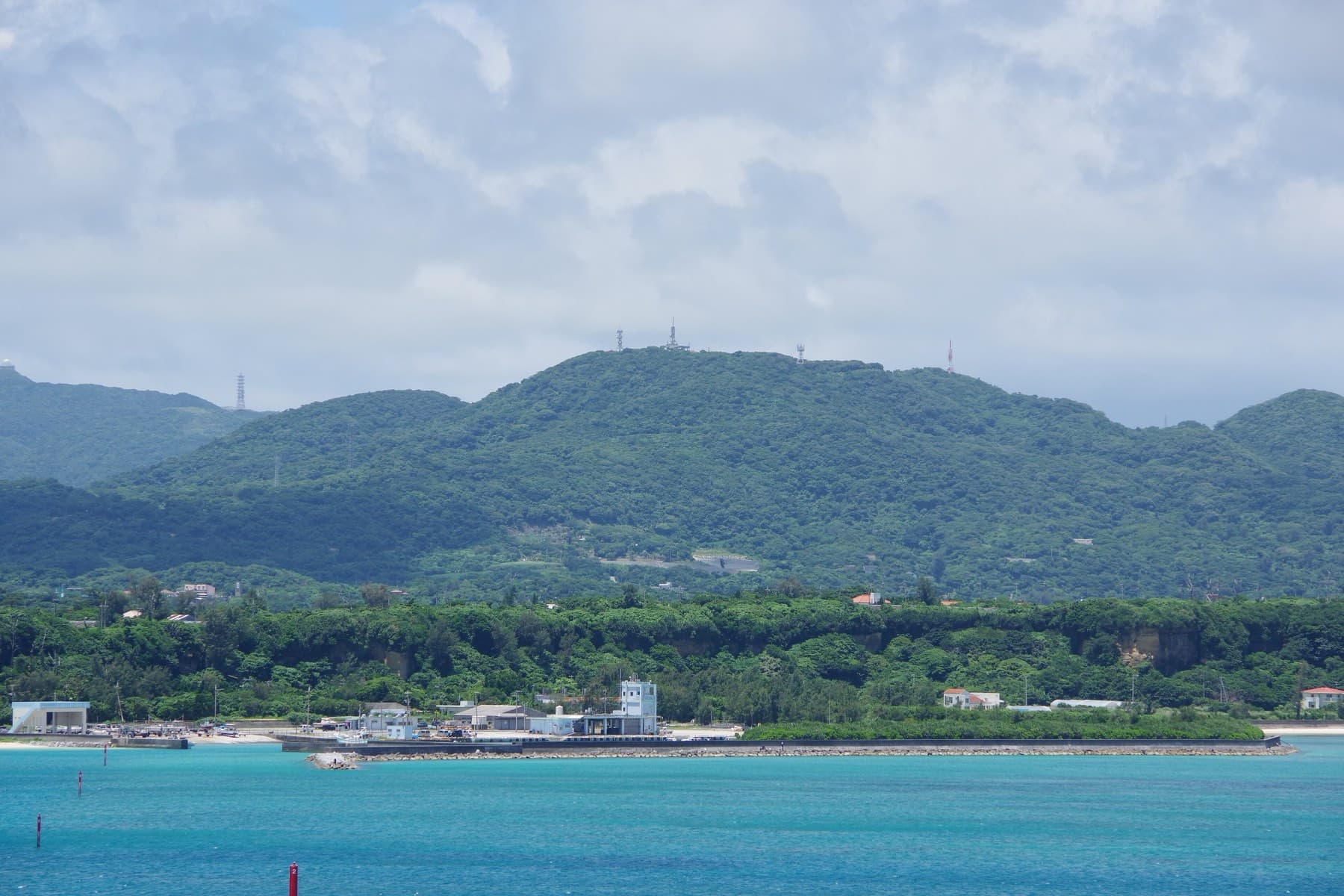 Across a blue channel from Kouri the forested ridge of Mount Oppa rises above a low shore of trees, a small harbour and white buildings