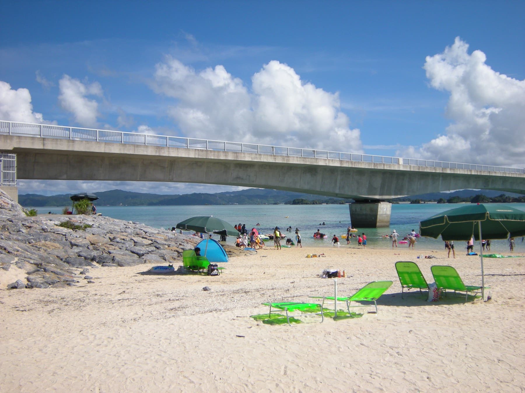 Green loungers and parasols dot the sand at Kouri Beach in August while swimmers fill the shallows under the concrete span of the Kouri Bridge