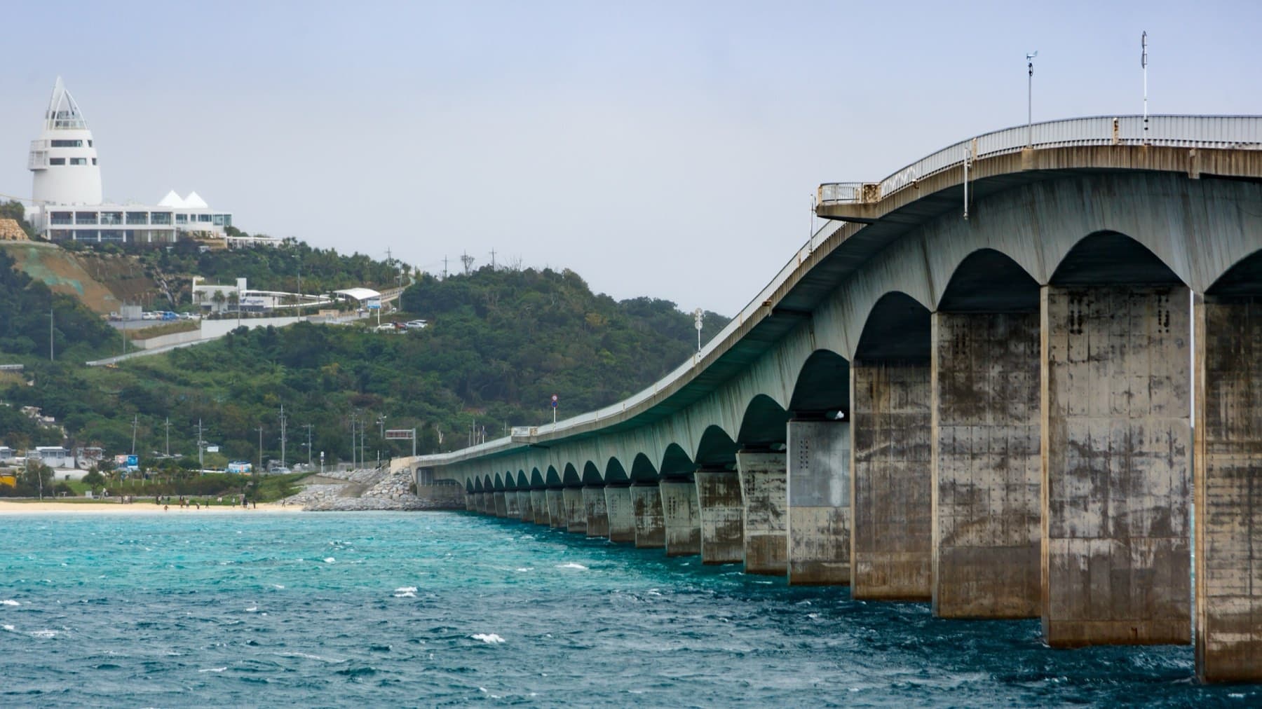 The concrete arches of the Kouri Bridge curve down to the Kouri shore over wind-ruffled blue water, with the white Kouri Ocean Tower on the hill above