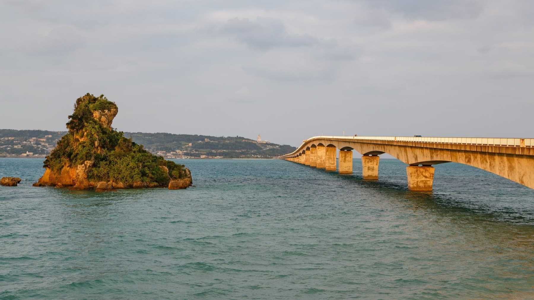 Low evening light turns a craggy rock islet and the piers of the Kouri Bridge gold as the bridge strides across the calm channel toward Yagaji