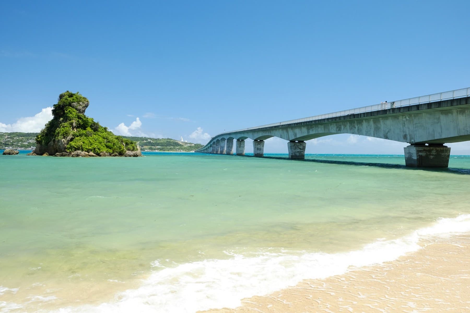 Small waves wash pale sand at the Yagaji end of the Kouri Bridge, where a rock islet capped with green stands in clear shallows beside the long span