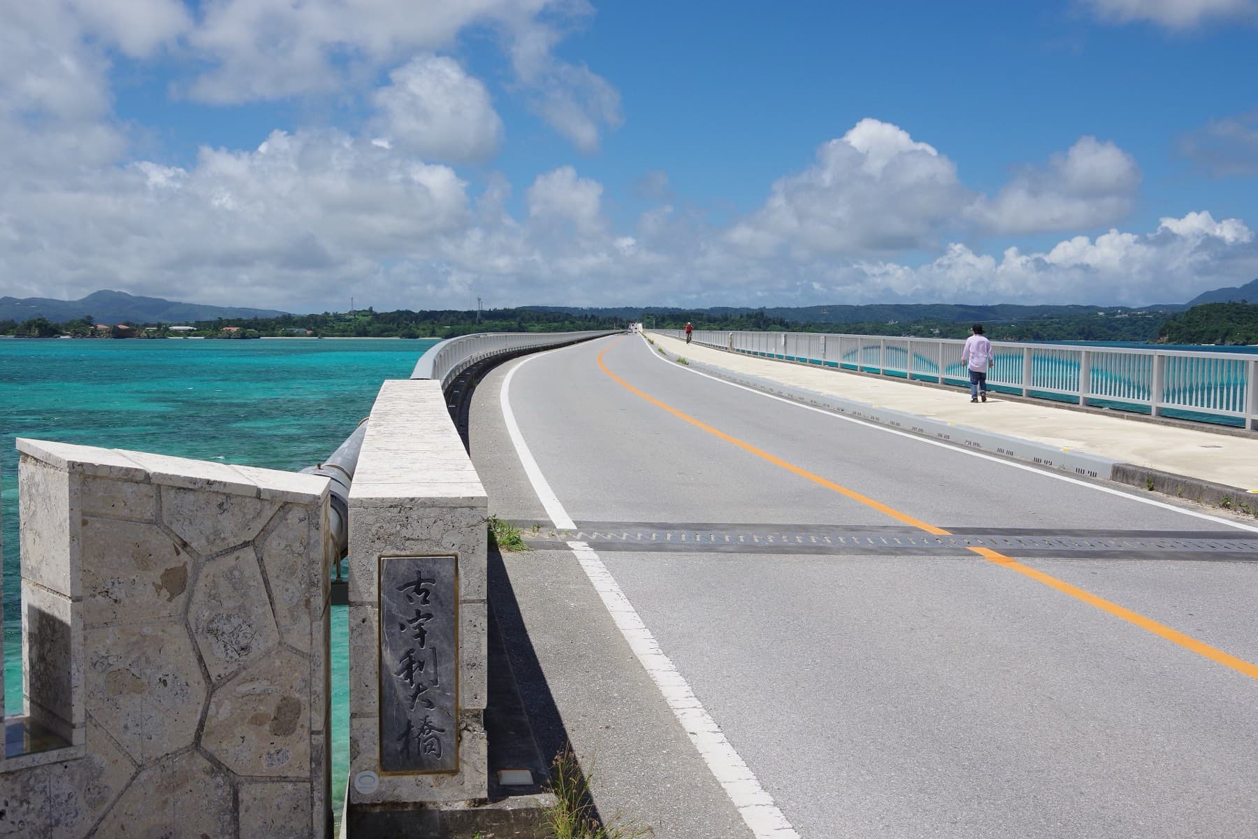 The two-lane deck of the Kouri Bridge curves out over pale green shallows from its carved stone name post, with a wide footway on the right and Yagaji's low hills behind.