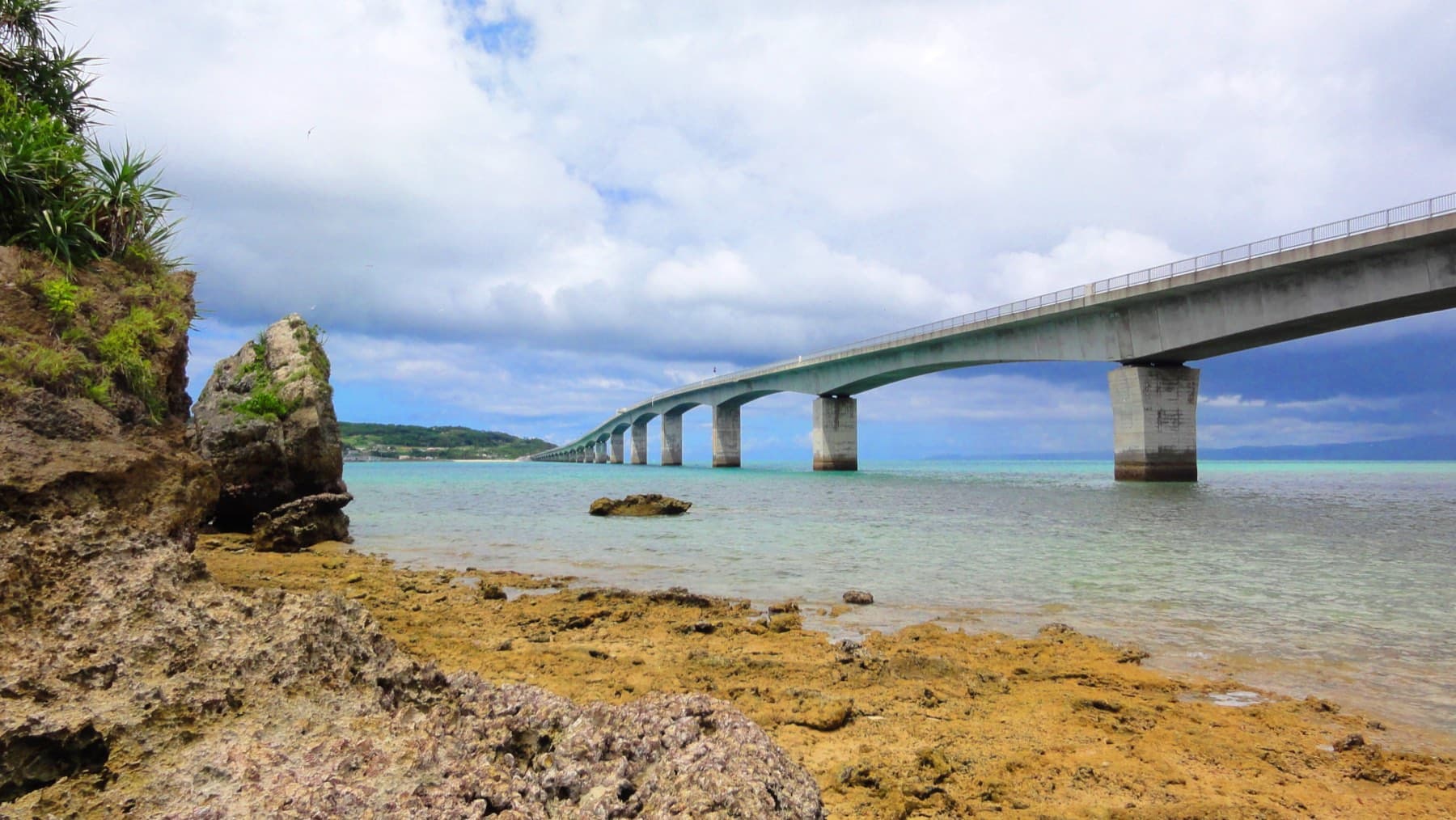 The long concrete Kouri Bridge strides across clear shallow water past a rocky coral shore and a small limestone islet under a bright summer sky.