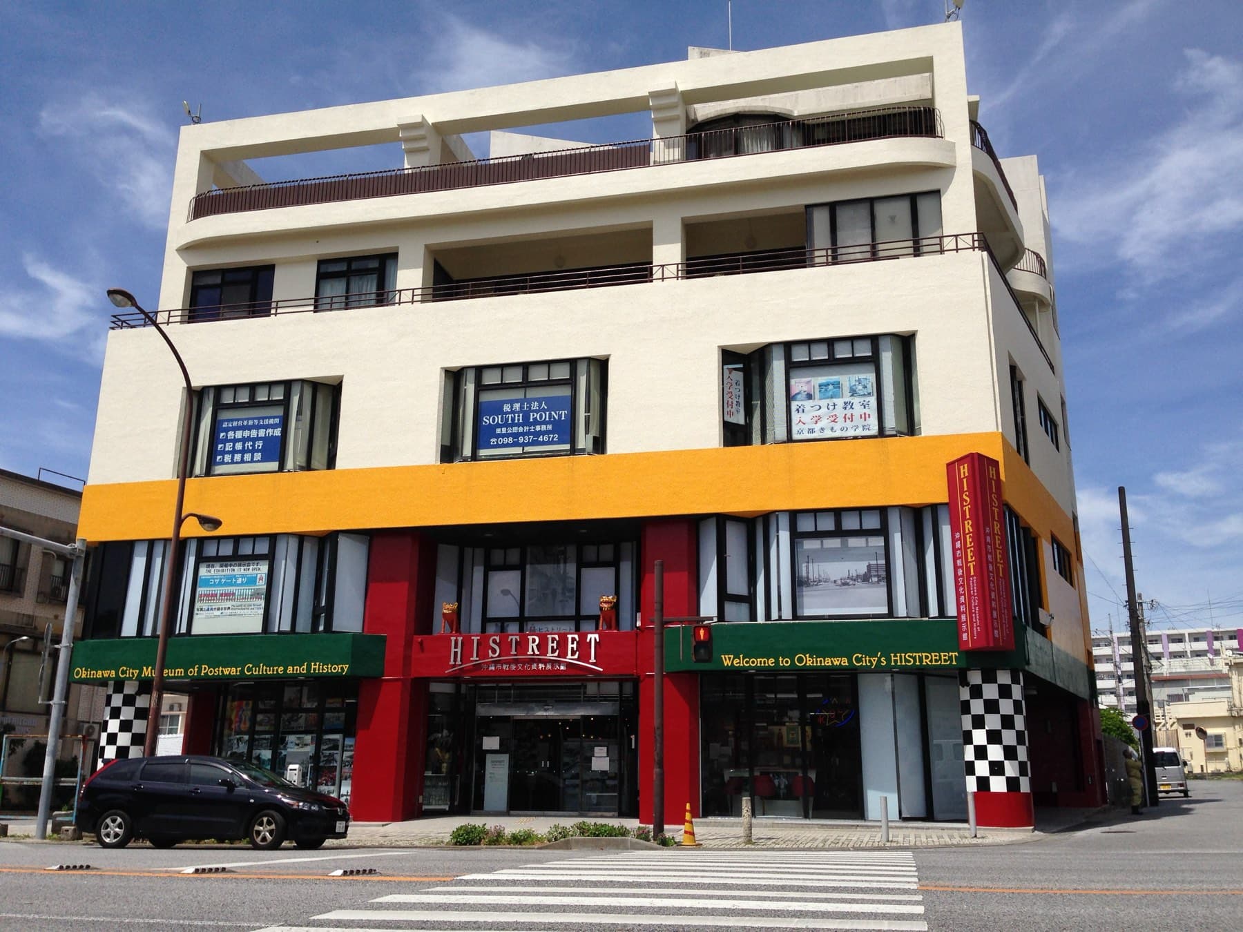 A corner building in Koza painted in red, orange and checkerboard trim houses the Histreet museum of postwar Okinawa City, under a blue sky with light cloud