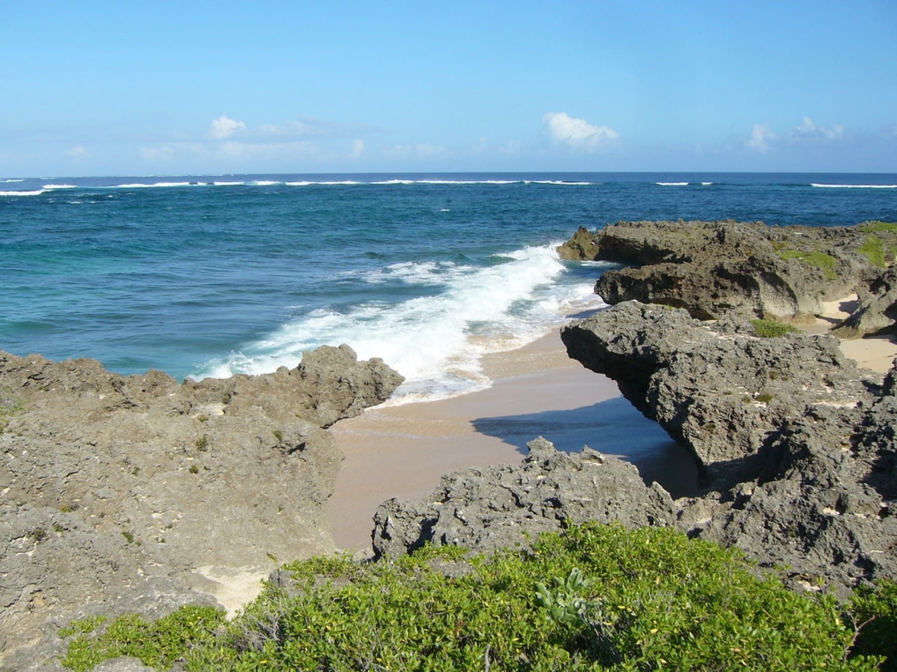 A small wave runs up a pocket of sand between rough limestone rocks on the Kudaka shore, with the reef line breaking white offshore