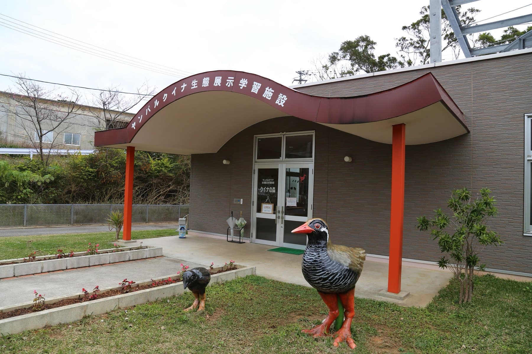 Two Okinawa rail statues stand on the lawn outside the curved entrance canopy of Kuina no Mori in Ada, Kunigami.