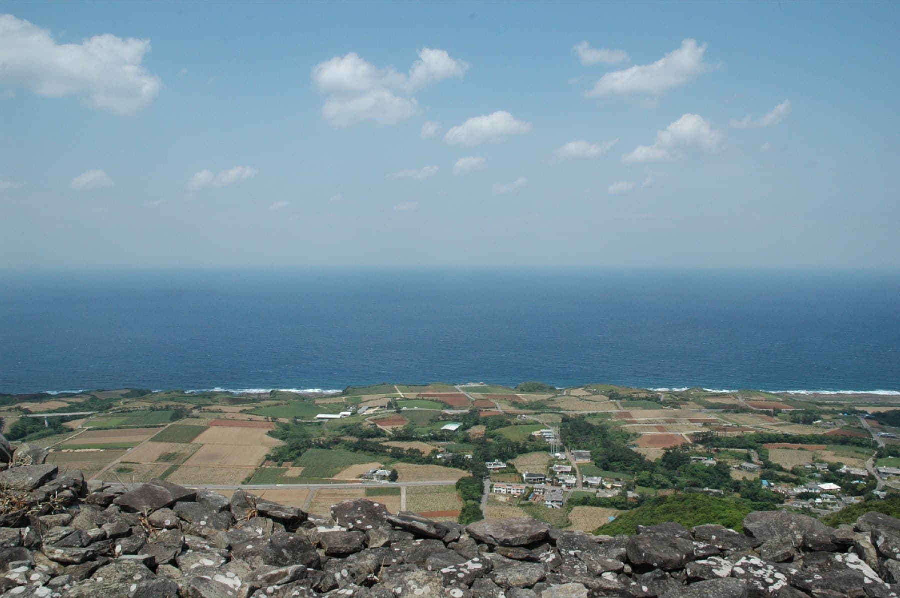 Patchwork farmland spreads from dark lava rocks on a hilltop on Kume-jima to a strip of surf and the deep blue East China Sea