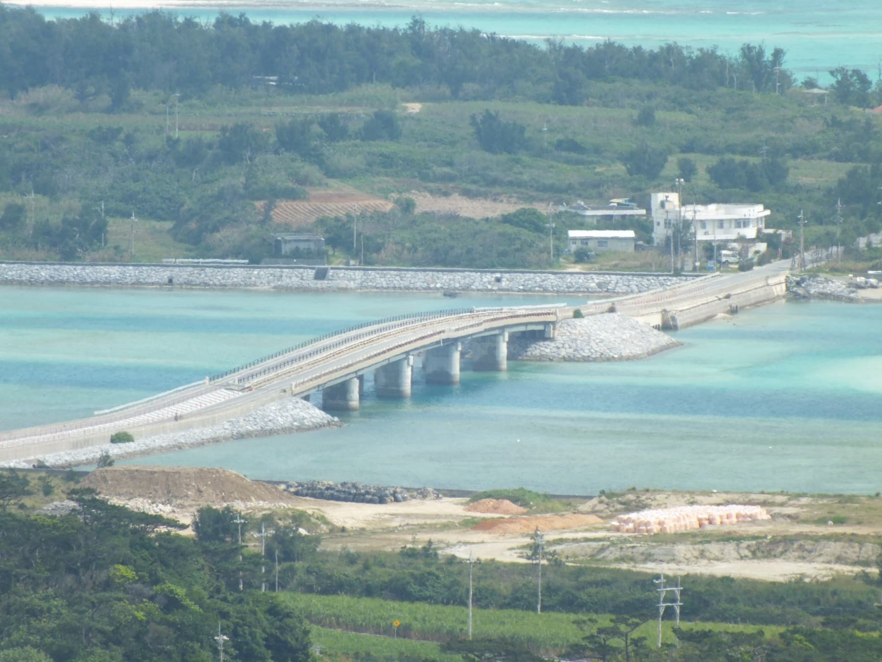 The low road bridge to Ou-jima crosses a pale green channel from Kume-jima, seen from the hills above with cane fields in the foreground and houses on the far shore.