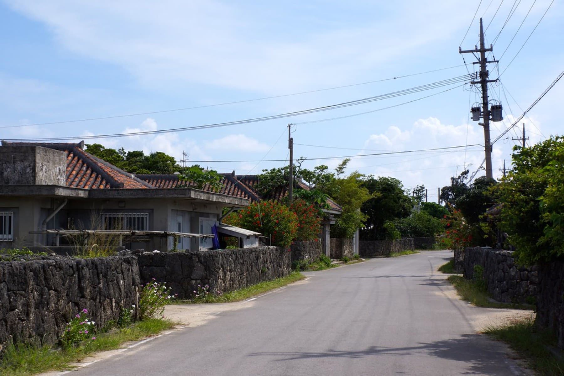 A quiet road bends past coral-stone walls and a red-tiled house in one of Kuroshima's hamlets.