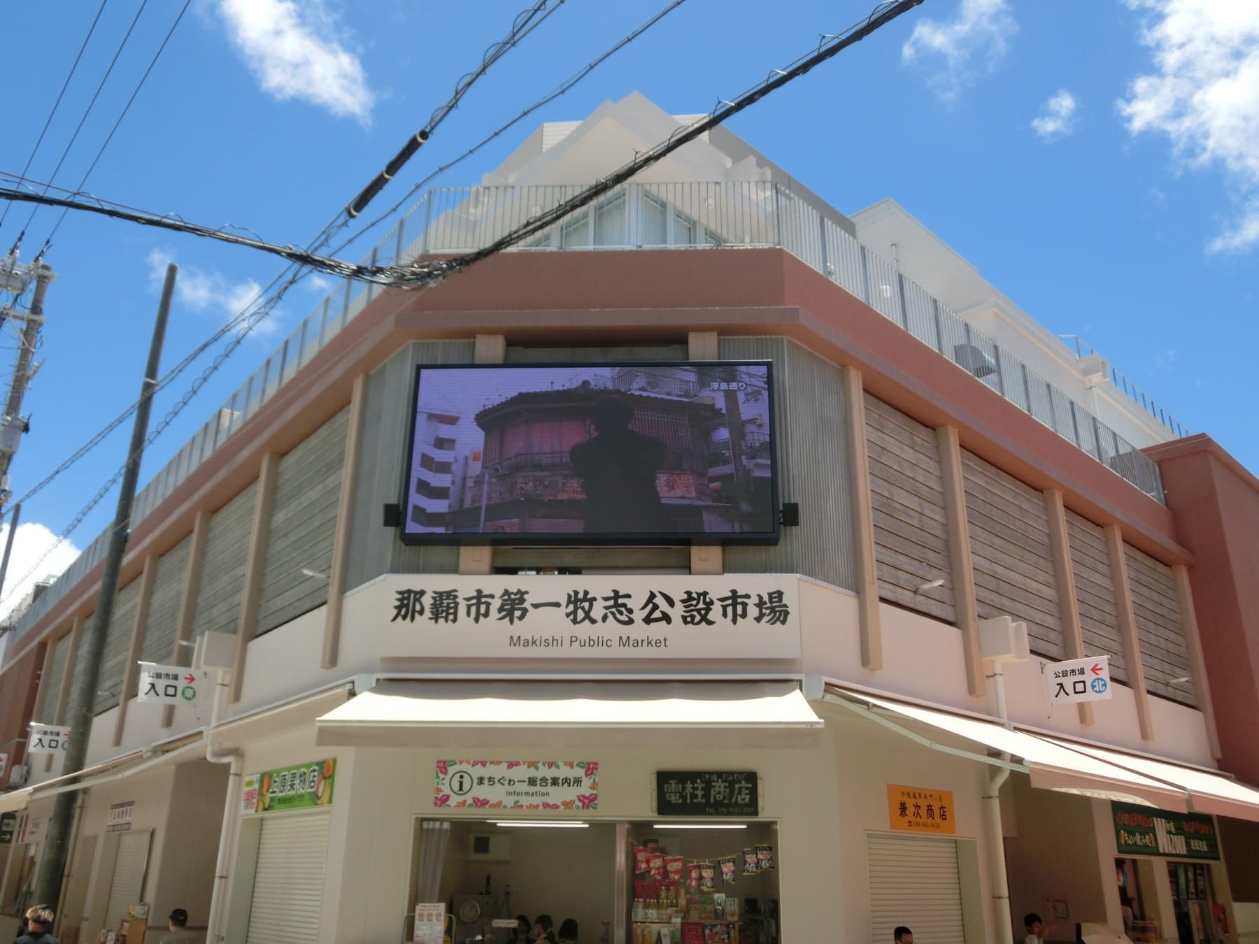The rebuilt Makishi First Public Market rises on a street corner in central Naha under a blue sky, and its name runs in Japanese and English above the entrance