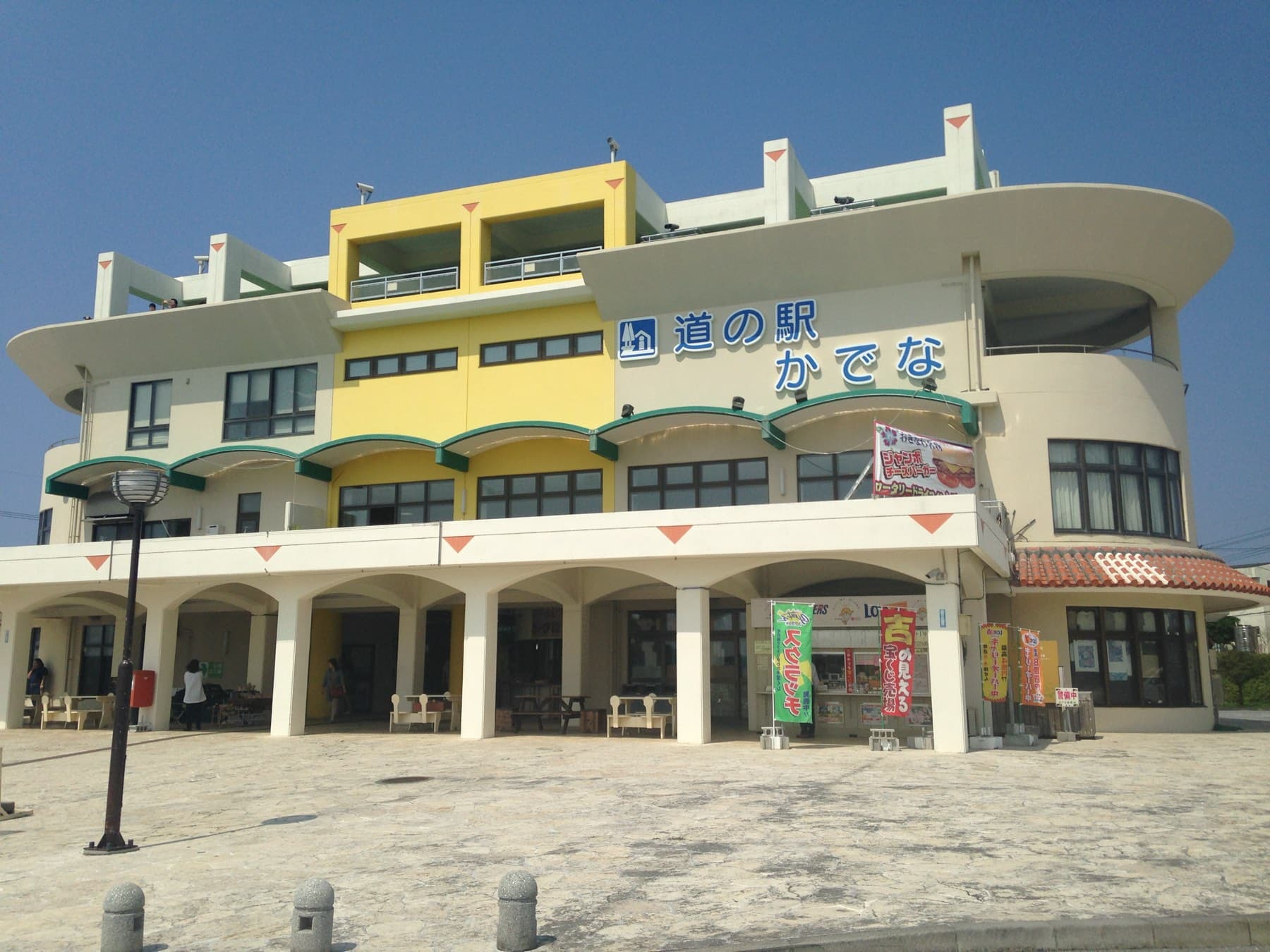 The curved cream and yellow building of Michi-no-eki Kadena stands behind a paved plaza with benches under its arches on a clear day