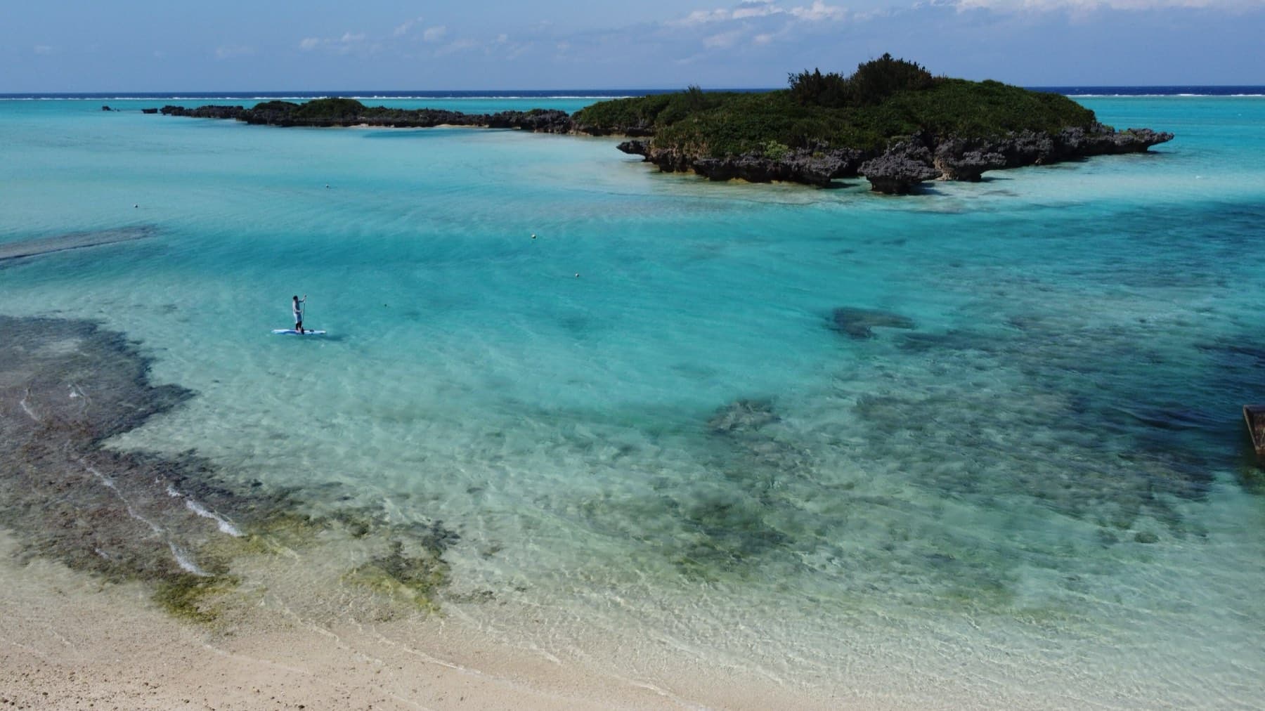 A wooded islet sits beyond the clear shallows of Minata Beach as a lone paddleboarder crosses the lagoon.