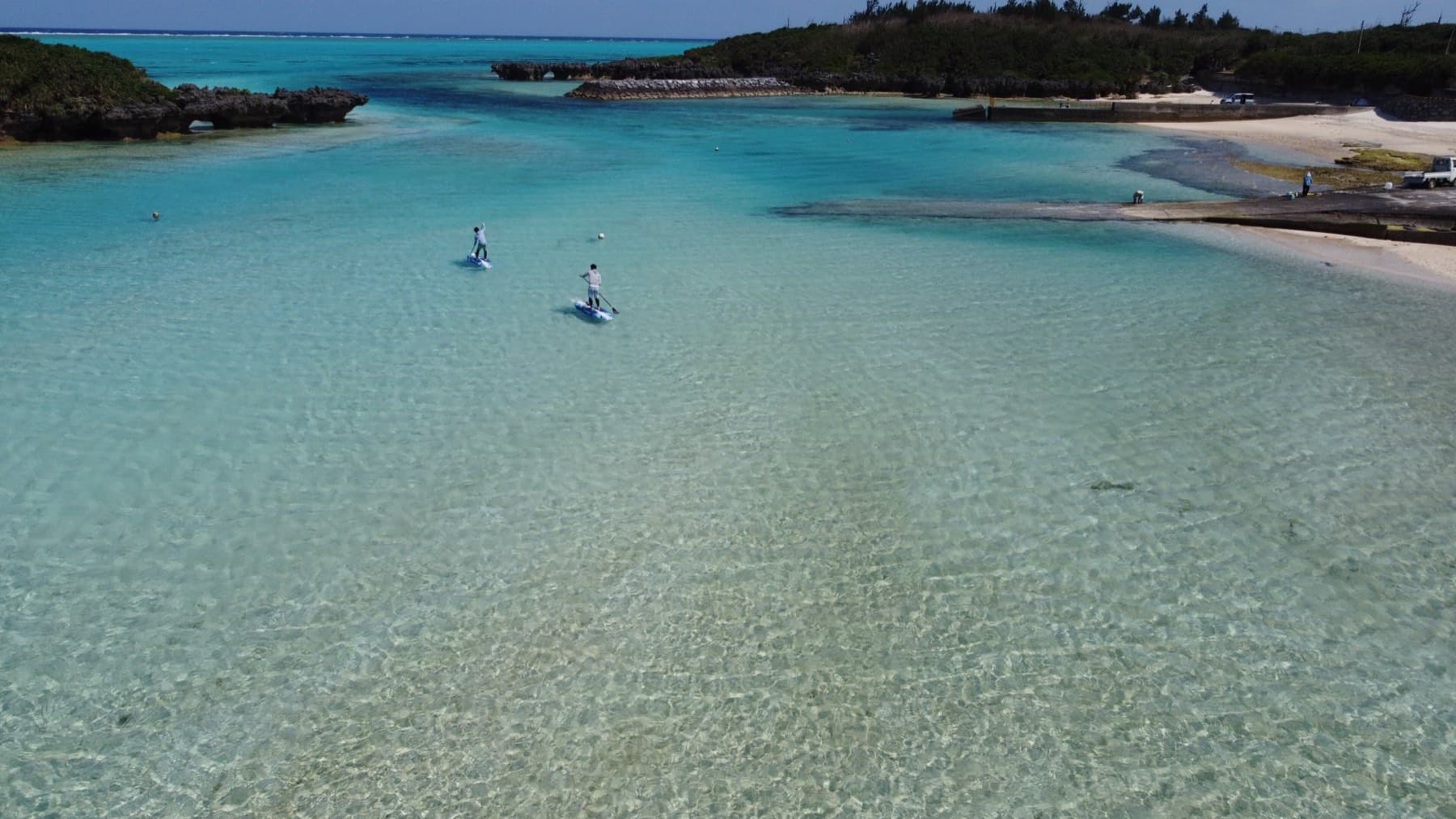 Two paddleboarders float on the clear green lagoon of Minata Beach, sheltered by low rocky headlands on Yoron.