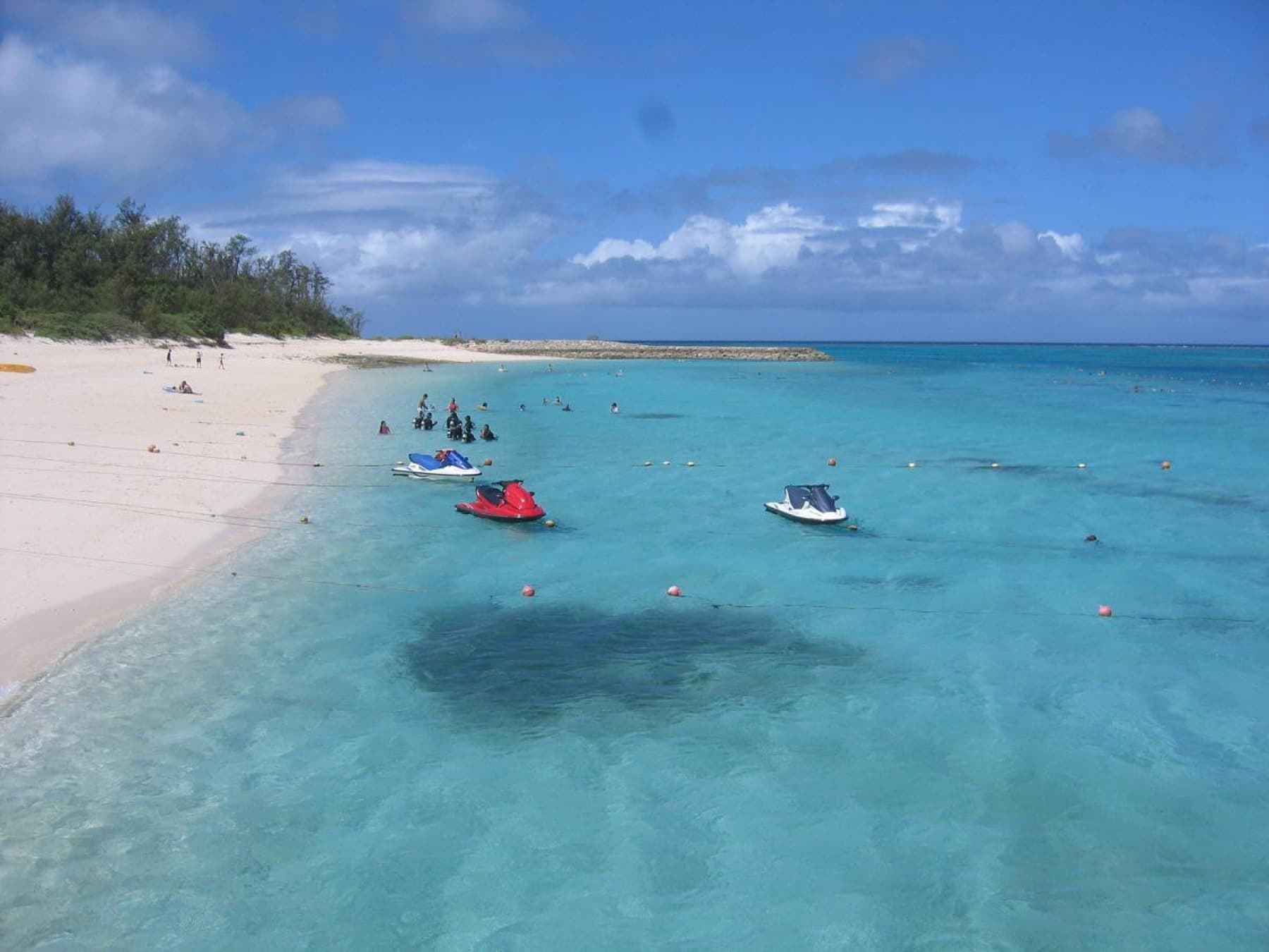 Jet skis and snorkellers float over a coral patch in the bright blue water off Minna Beach, where white sand curves away below dense trees