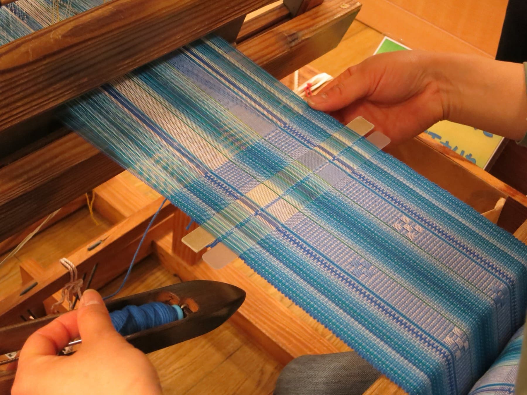 Hands guide a dark shuttle wound with blue thread across a minsah cloth of pale blue stripes and woven squares on a wooden loom on Ishigaki