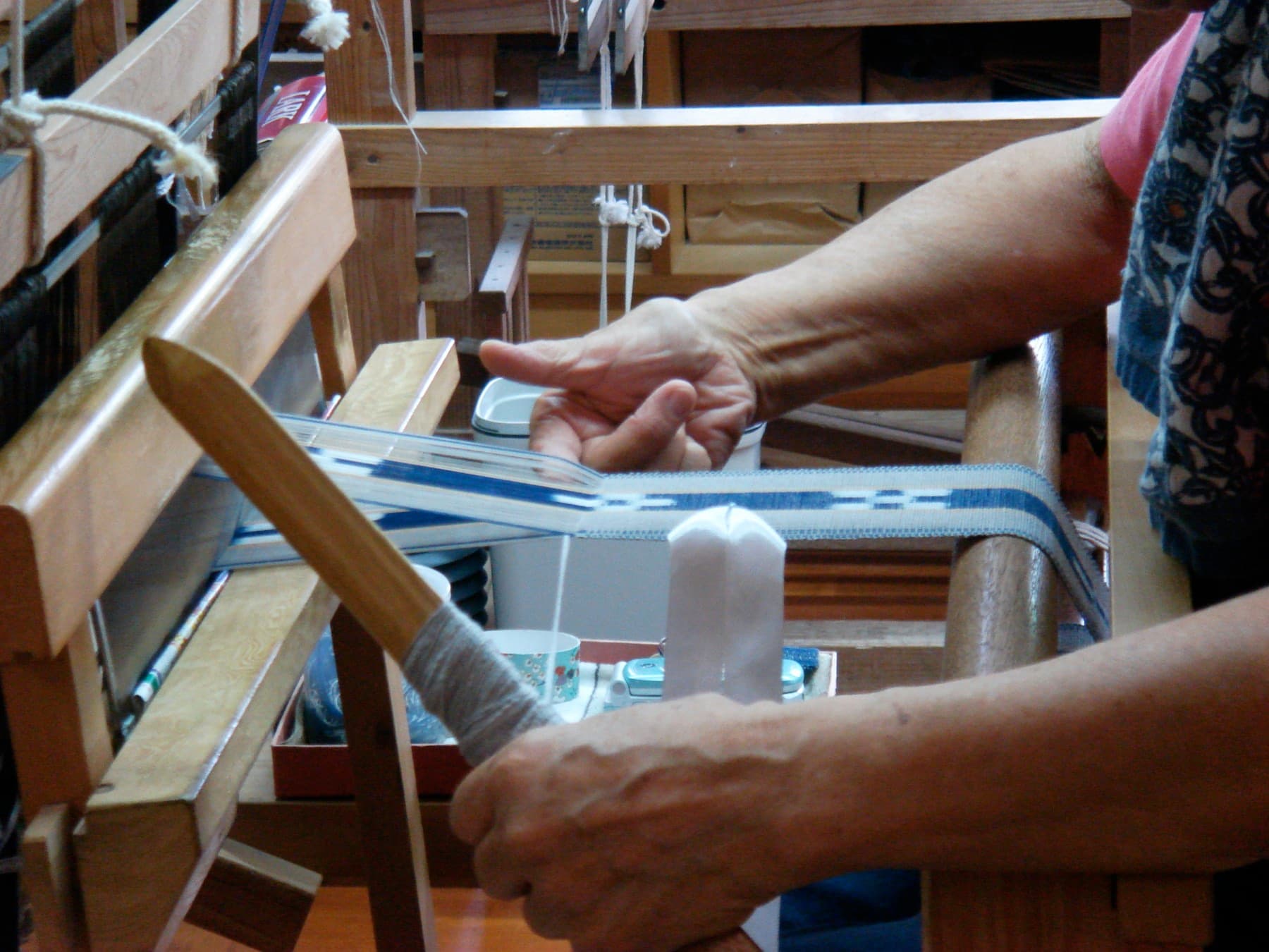 A weaver's hands pass a wooden shuttle through a narrow blue and white minsah belt on a hand loom in a workshop on Taketomi