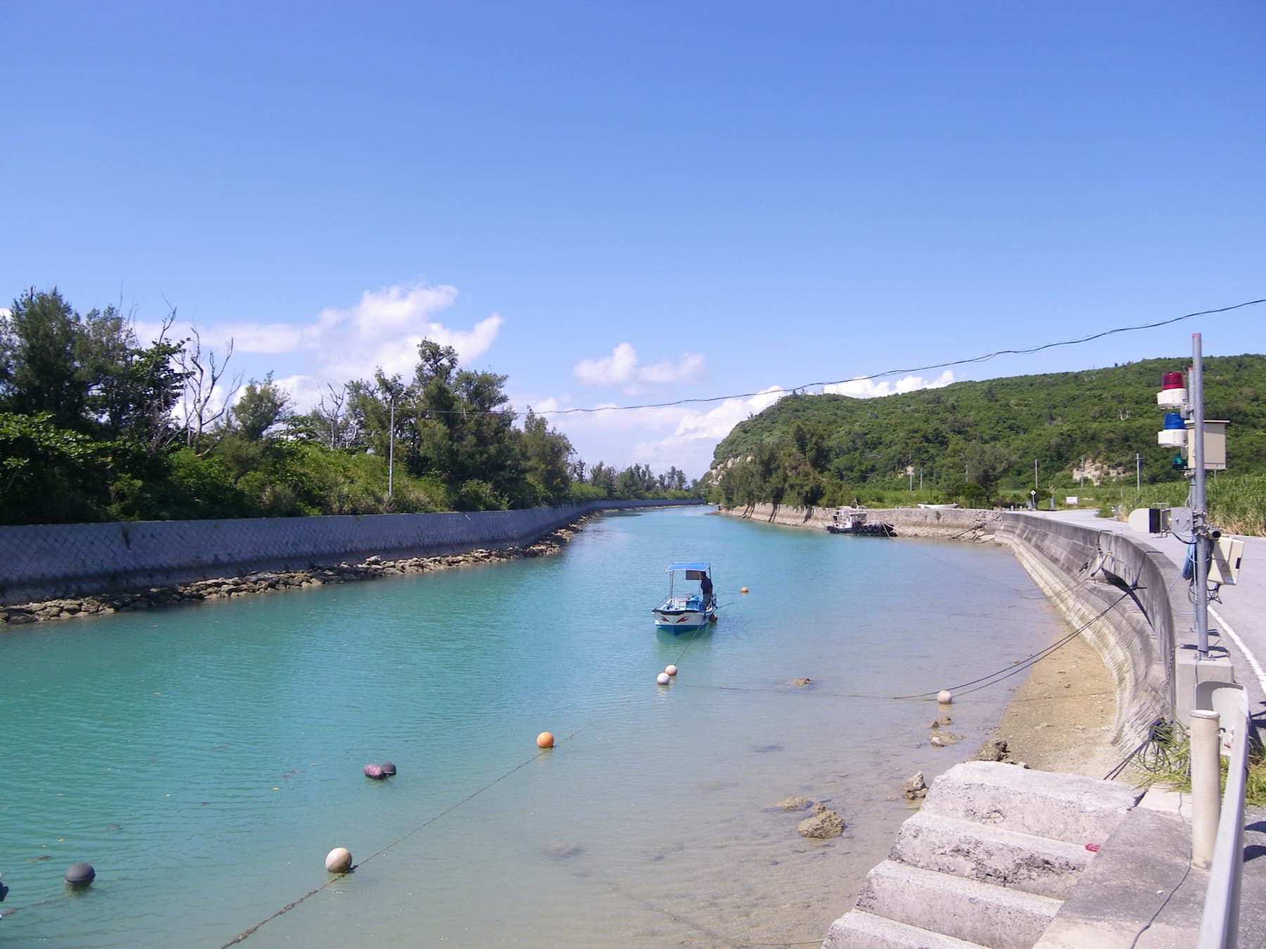 A small fishing boat floats in the narrow green channel between Miyagi and Henza, with seawalls on both banks and wooded hills under a summer sky
