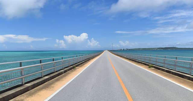 A wide view along the deck of the Irabu Bridge, with its orange centre line, broad shoulders and low railings running straight out between blue sea on both sides.