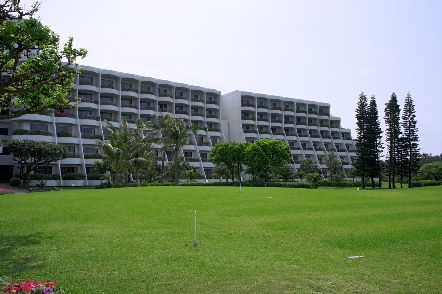 A wide green lawn and tall Norfolk pines stand in front of the terraced white block of the Miyakojima Tokyu Hotel and Resorts.