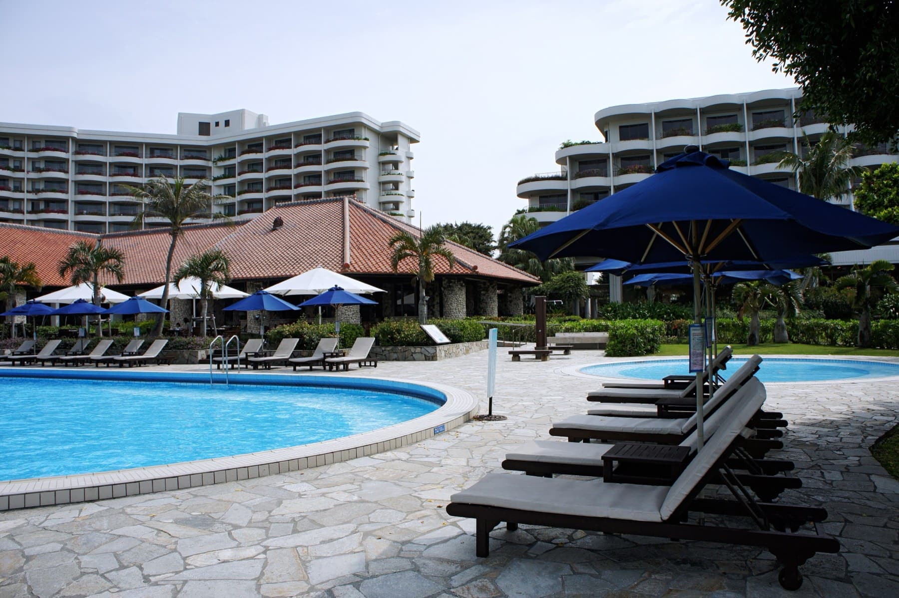 Blue umbrellas shade sun loungers around the curving pool of the Miyakojima Tokyu Hotel and Resorts in front of its low white wings.