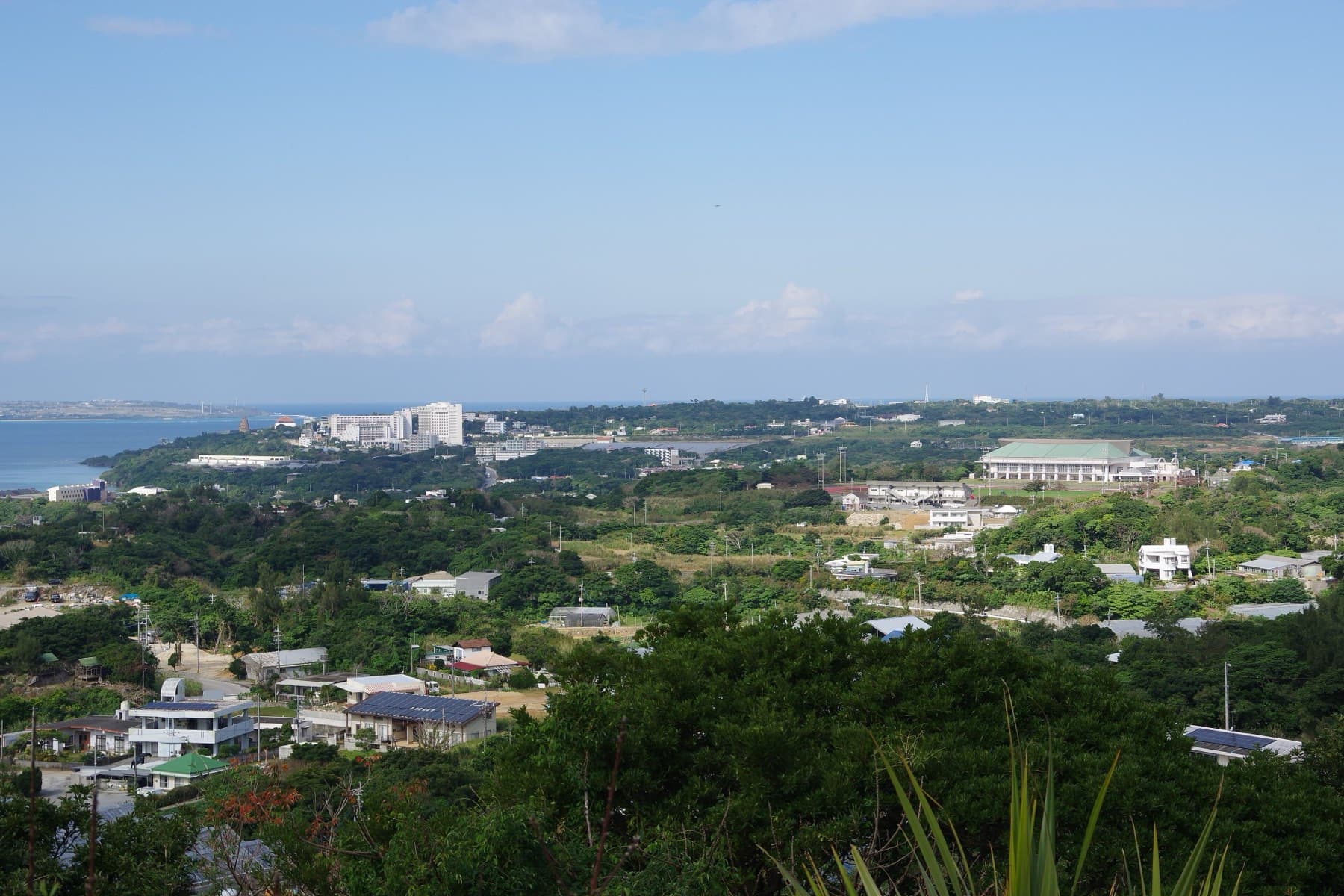 Green hills and scattered houses of northern Motobu roll down toward the coast and Ocean Expo Park, with a pale sea and Ie-jima on the horizon.
