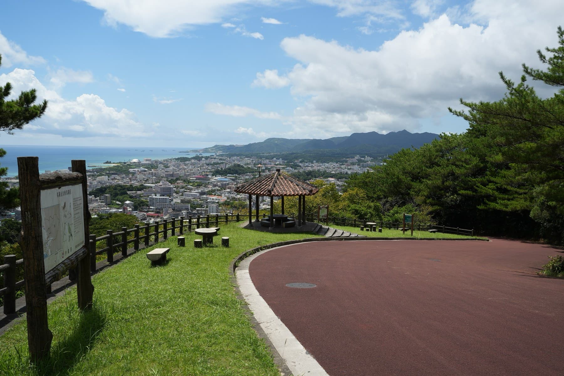 From a lookout in Nago Central Park the city spreads along Nago Bay below green hills, with a gazebo beside a curving red path in the foreground