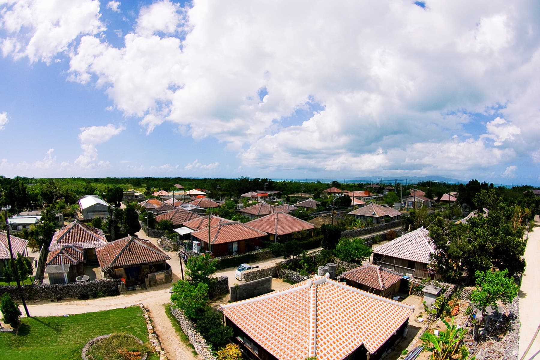 Red-tiled roofs and coral-stone walls fill the view over Taketomi village from the top of Nagomi-no-to on a day of big white clouds.