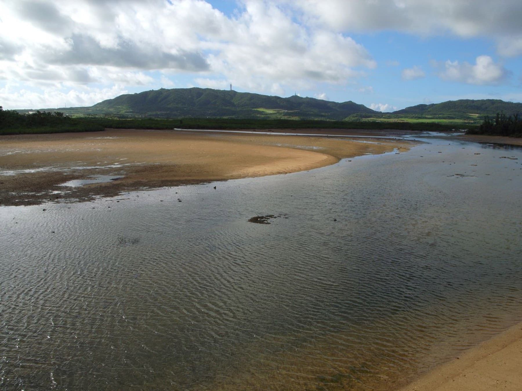 Low tide leaves rippled sand and shallow channels across the Nagura Anparu wetland under green hills on the west side of Ishigaki.