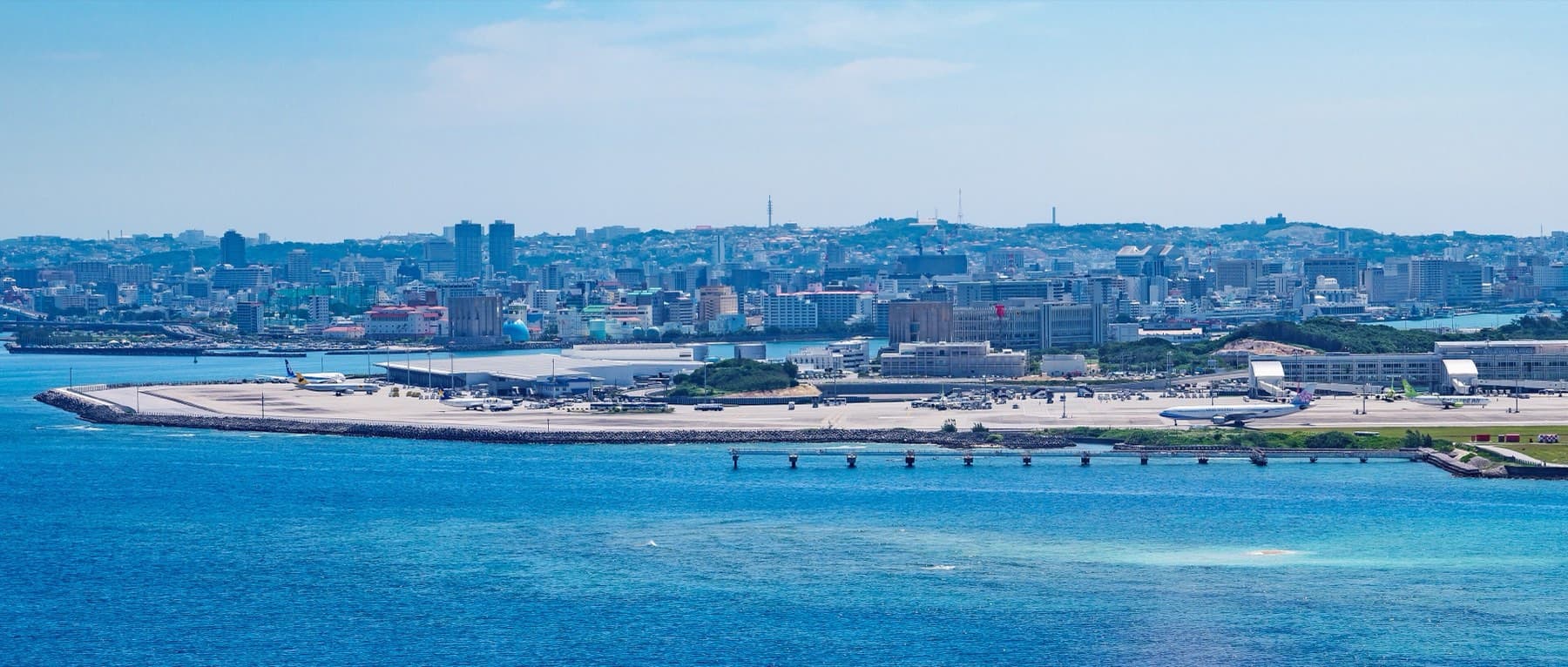 Blue water fills the foreground below Naha Airport, where planes stand at the terminal and the low city of Naha spreads along the shore behind