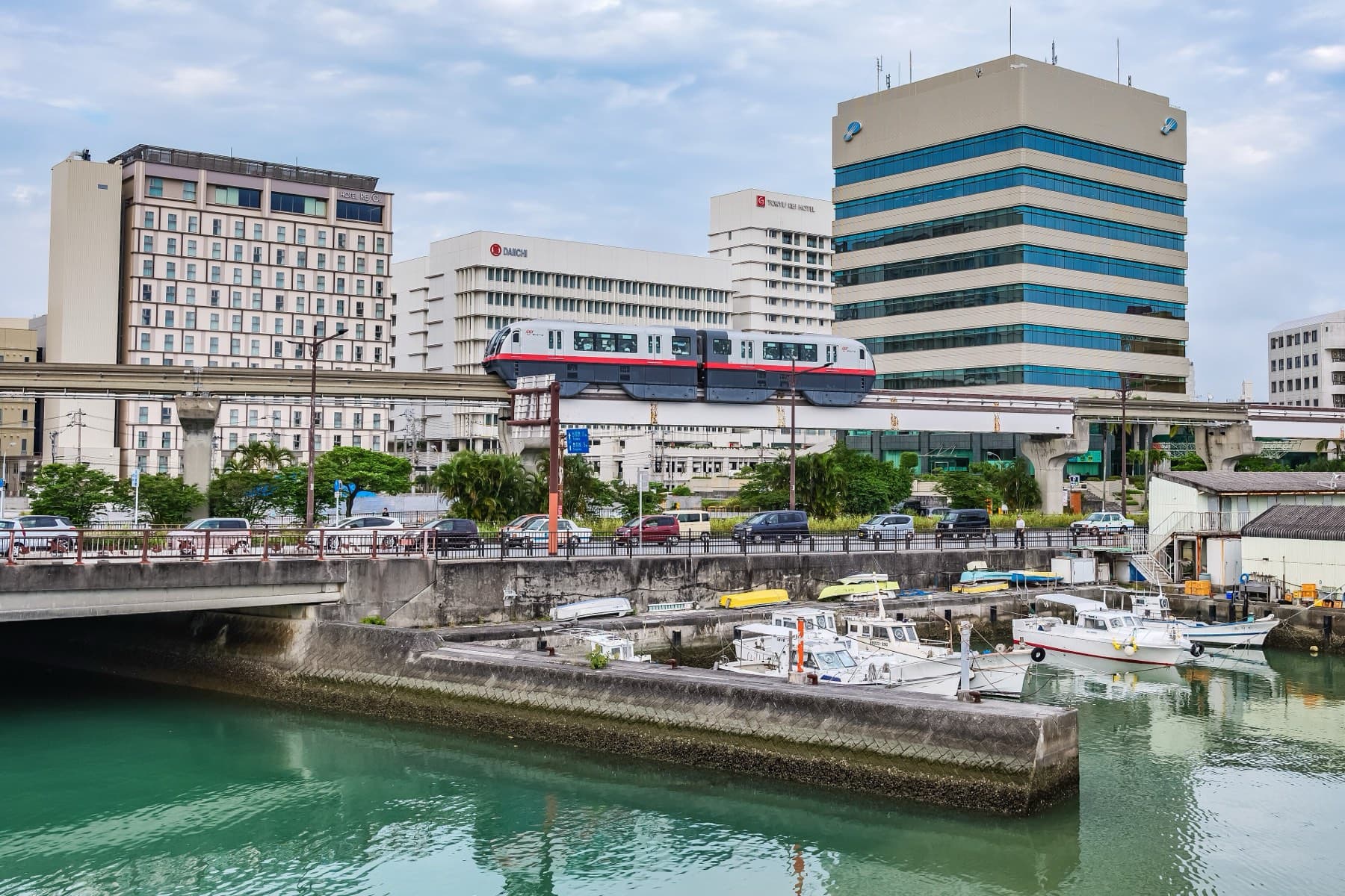 A Yui Rail train crosses a bridge above the Kokuba River in Naha, where small boats sit moored along the green water below office blocks