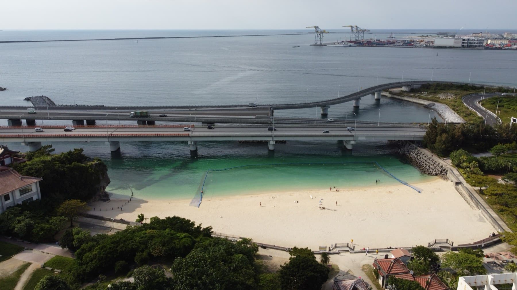 An aerial view of Naminoue Beach in Naha, a small sheltered cove of pale sand and green water tucked under the curving bridges of the coastal bypass.