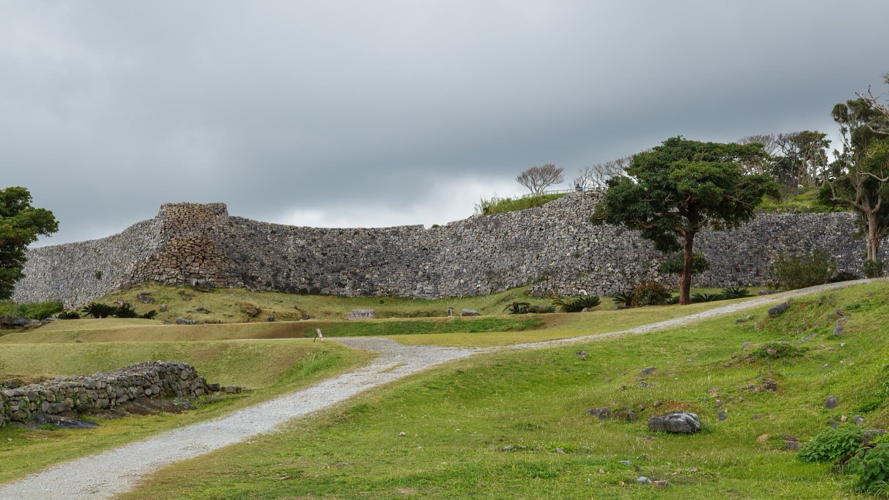 A gravel path curves across a lawn toward the high outer wall of Nakijin Castle under a heavy grey sky