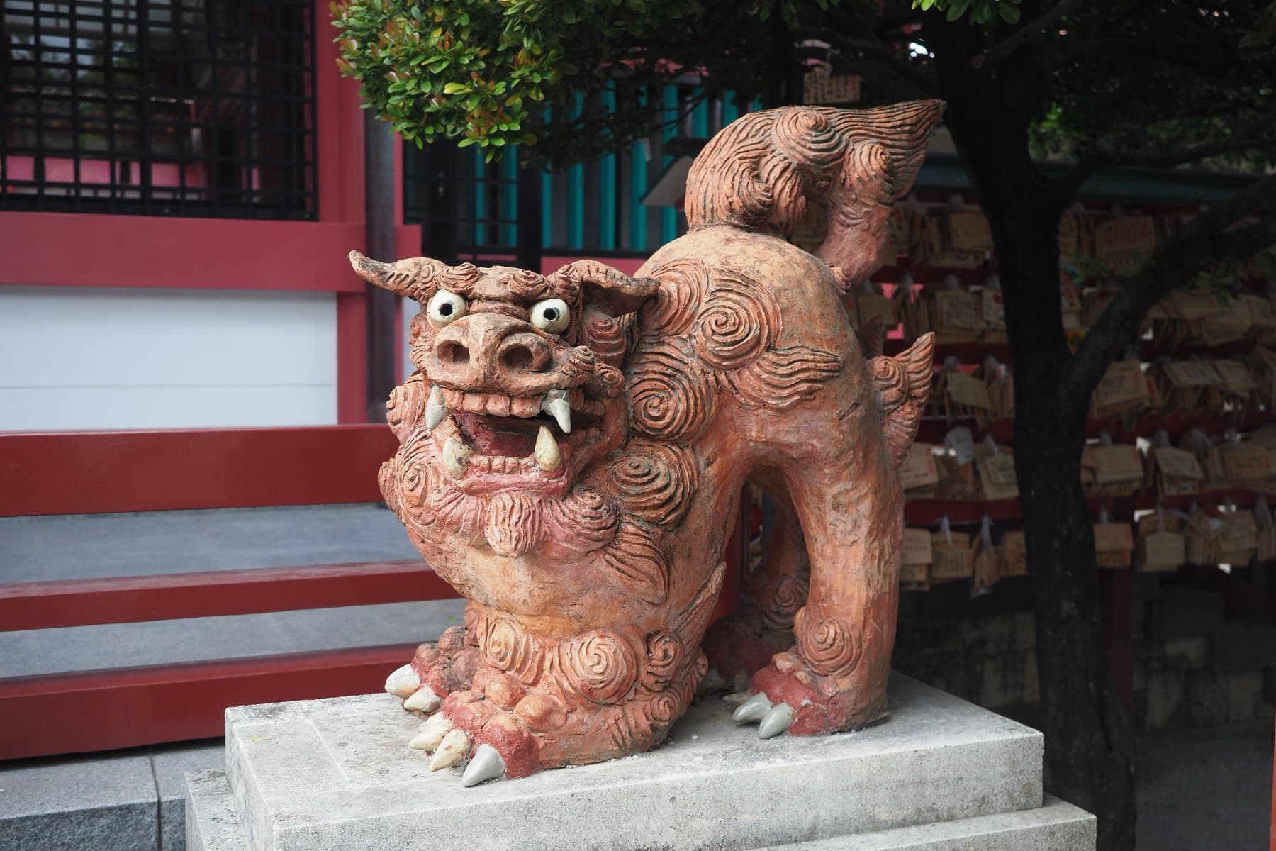 A curly-maned shisa of red clay bares its teeth on a stone plinth at Naminoue Shrine while wooden prayer plaques hang on a rack behind it