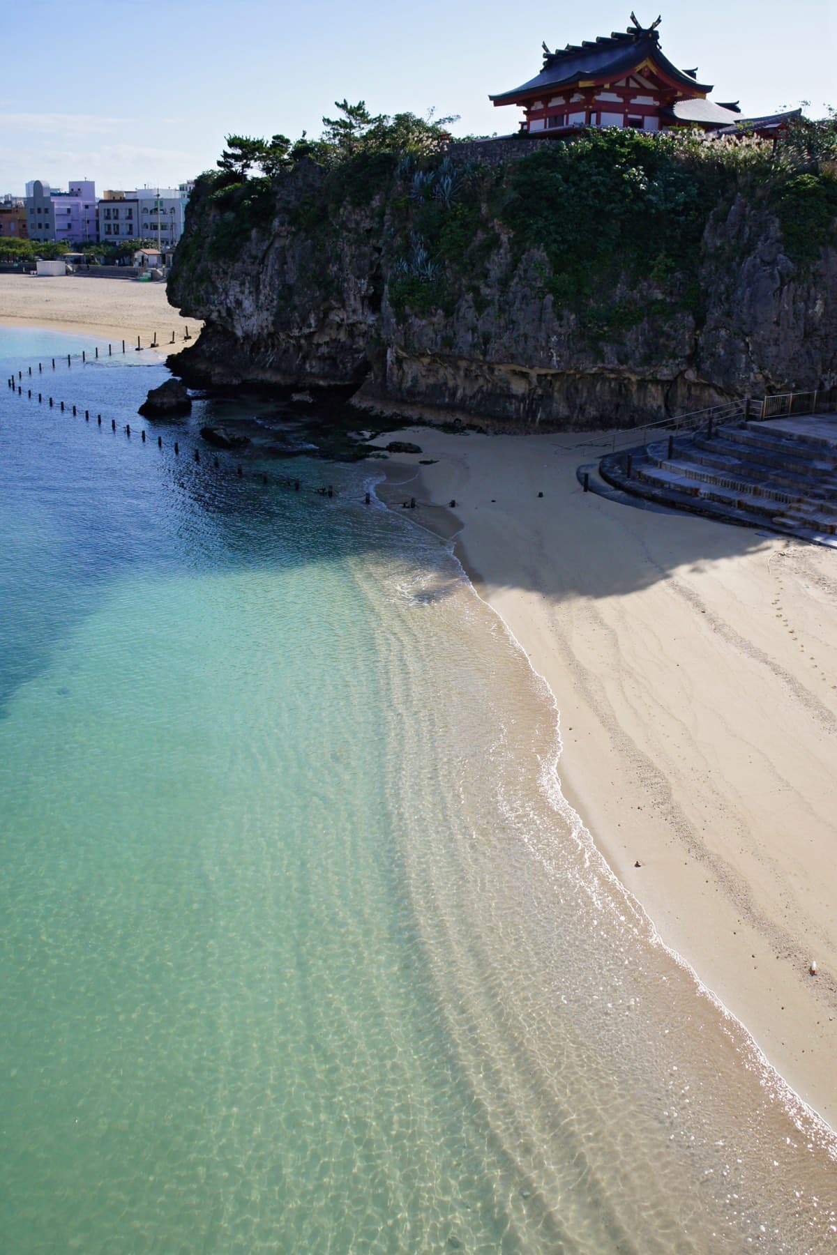 Clear green water laps the curve of Naminoue Beach below a limestone cliff where the red and black roof of Naminoue Shrine looks out to sea