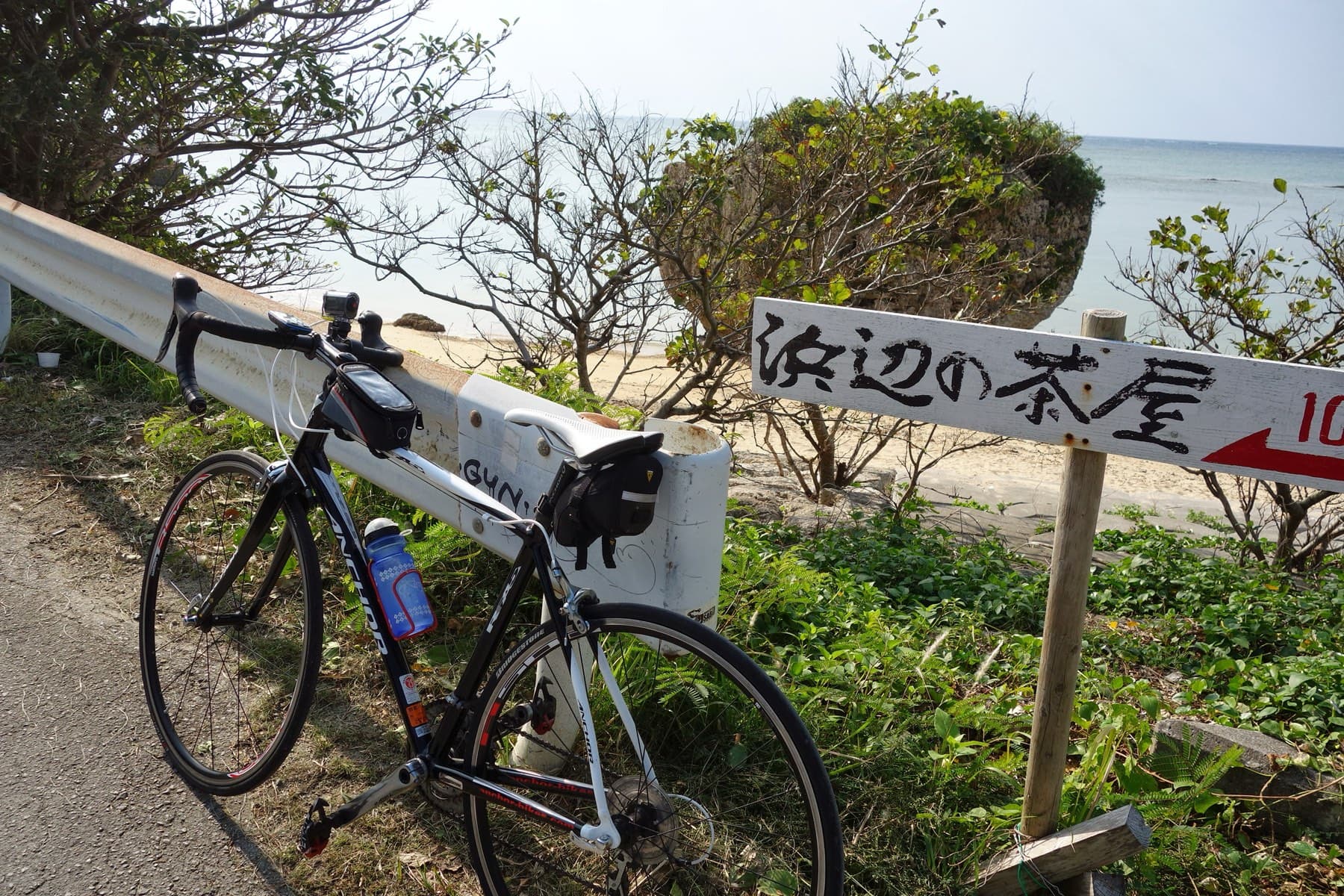 A road bike leans on a white guardrail beside a wooden sign for Hamabe no Chaya at the western end of Niibaru Beach, with scrub and the sea behind.