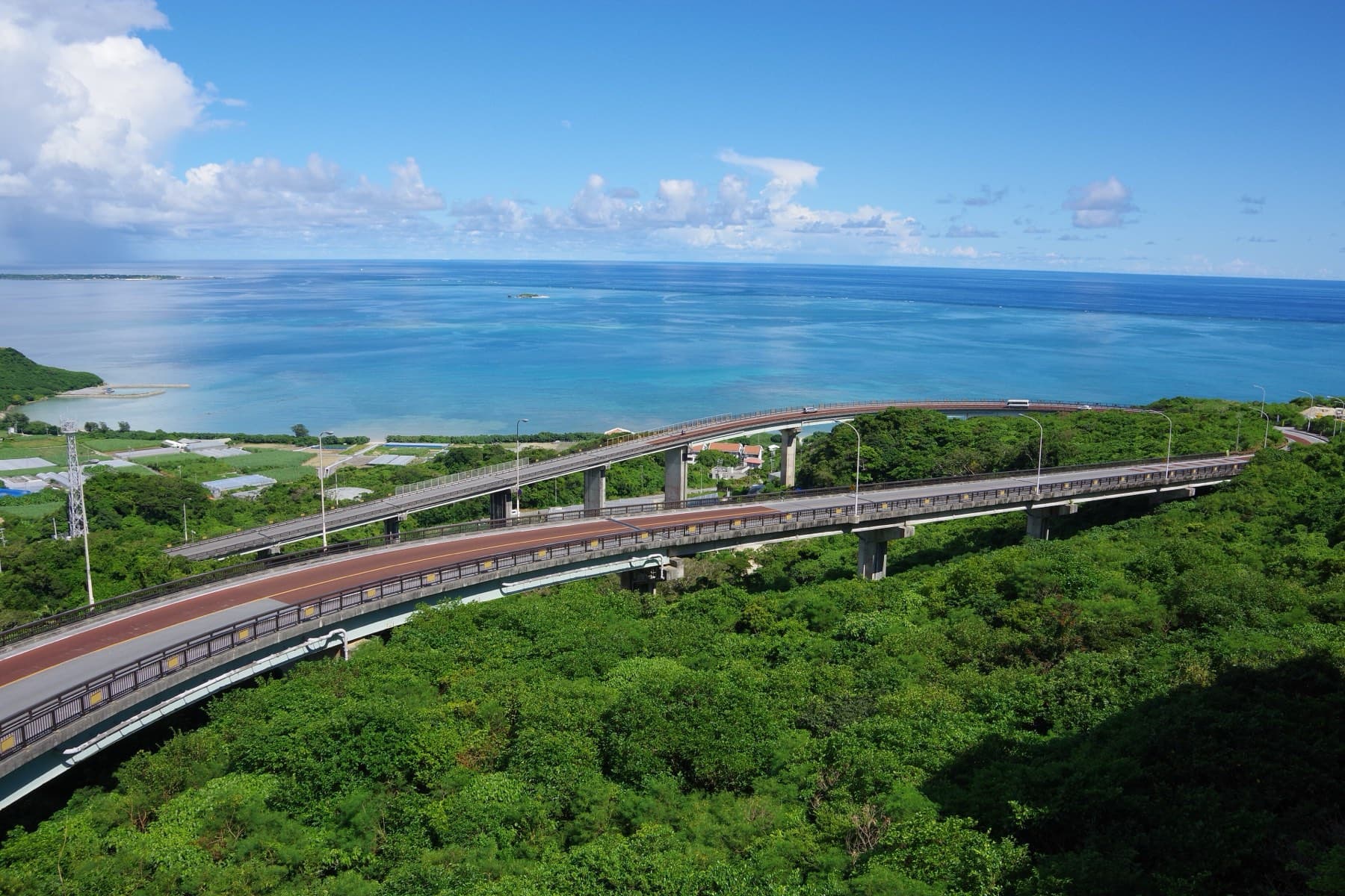 The double hairpin of the Niraikanai Bridge sweeps down through green forest toward the coast at Chinen, with the wide blue Pacific beyond