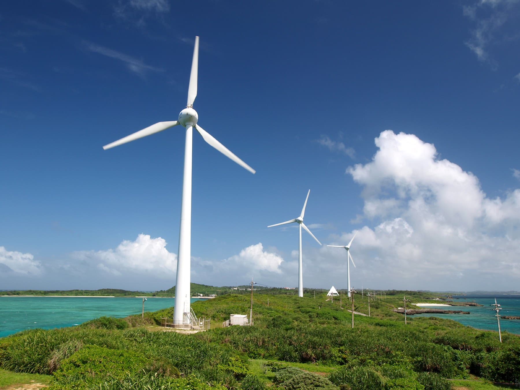 Two white wind turbines turn above green scrub at Nishi-Hennazaki on Miyako's northern tip while Ikema sits low on the sea beyond.
