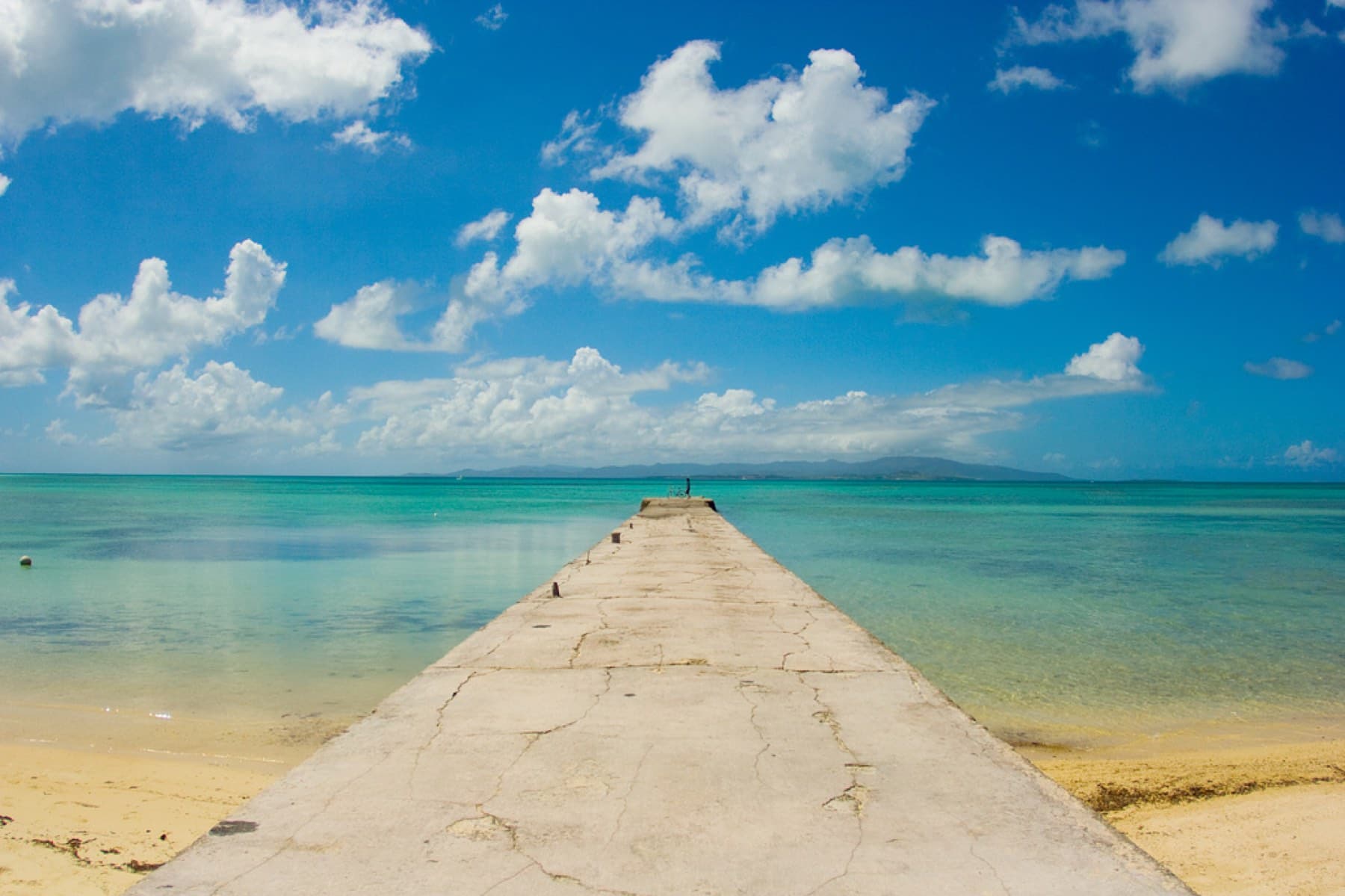 The concrete Nishi Pier runs straight out from the beach into pale green water on the west side of Taketomi under puffy white cloud.