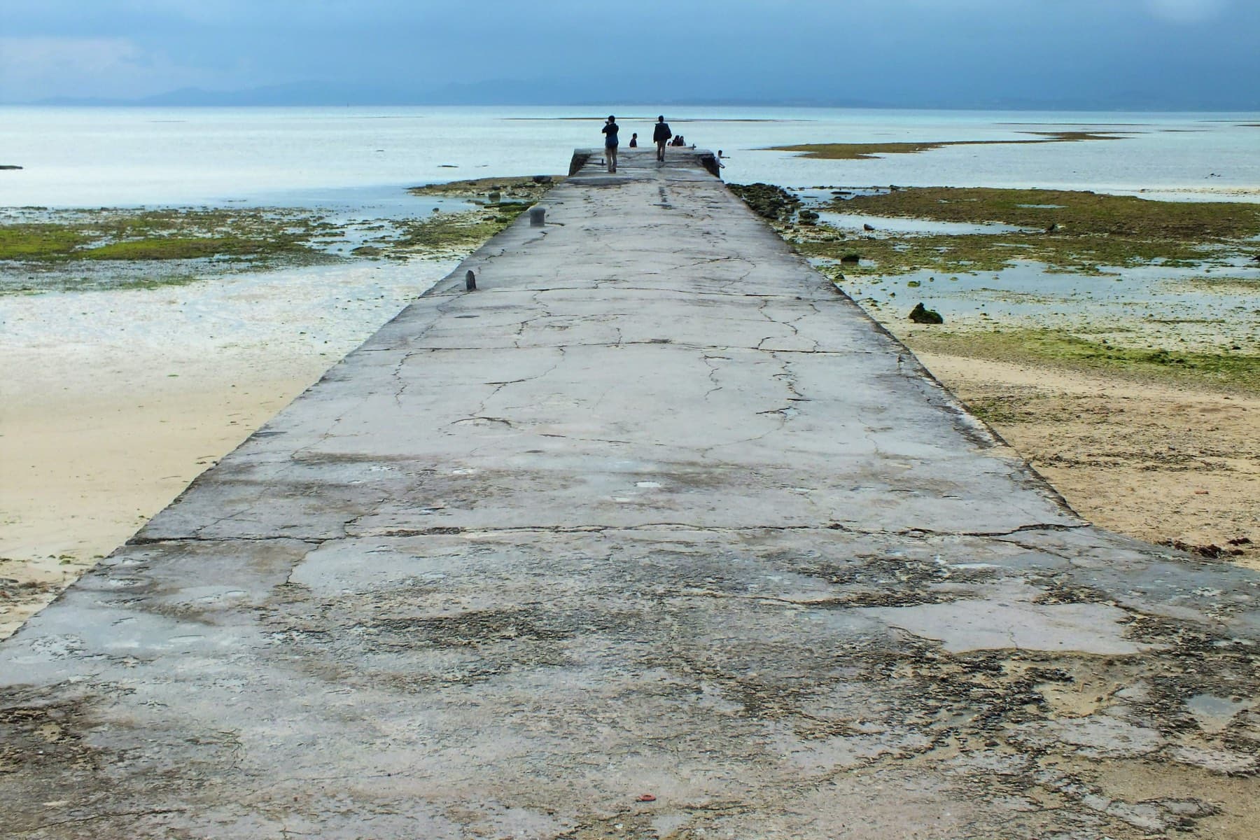 Low tide exposes weed-covered reef on both sides of the cracked concrete Nishi Pier on Taketomi as two figures stand at its far end.