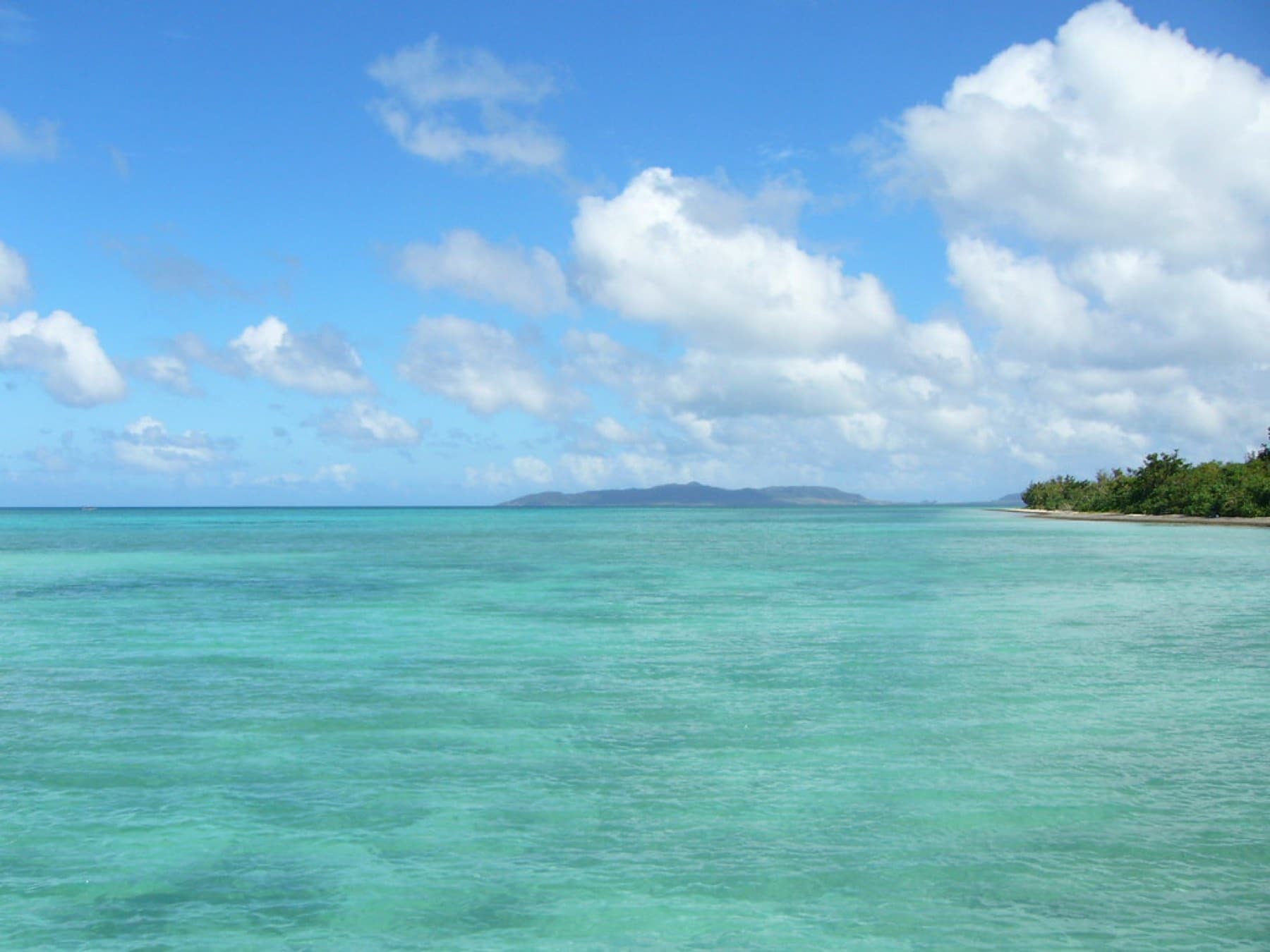 Shallow turquoise water stretches west from the Nishi Pier on Taketomi toward the low outline of a neighbouring island on the horizon.