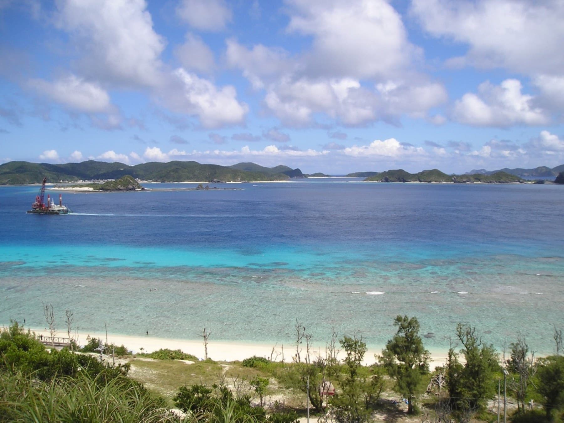 Nishibama Beach on Aka runs white below casuarina trees while the reef spreads pale green into deep blue water toward the hills of Zamami