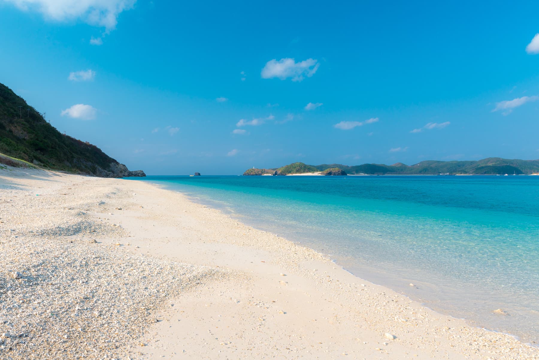 Coarse coral sand slopes into clear pale water along Nishibama Beach on Aka, and low wooded islands line the far side of the channel