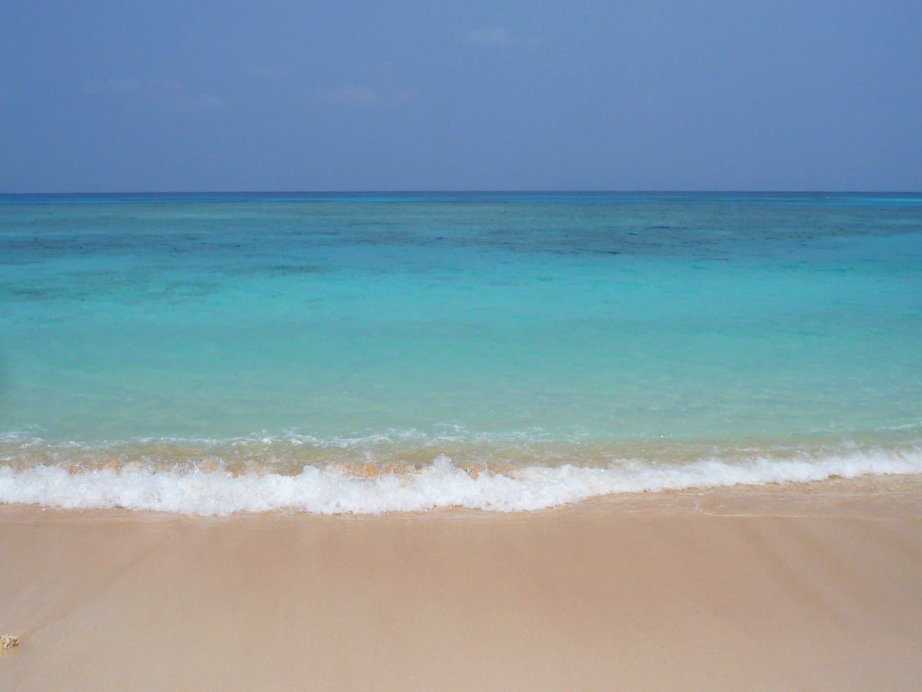 A thin line of surf breaks on the pale sand of Nishihama as the water shades from clear aqua to deep blue offshore.