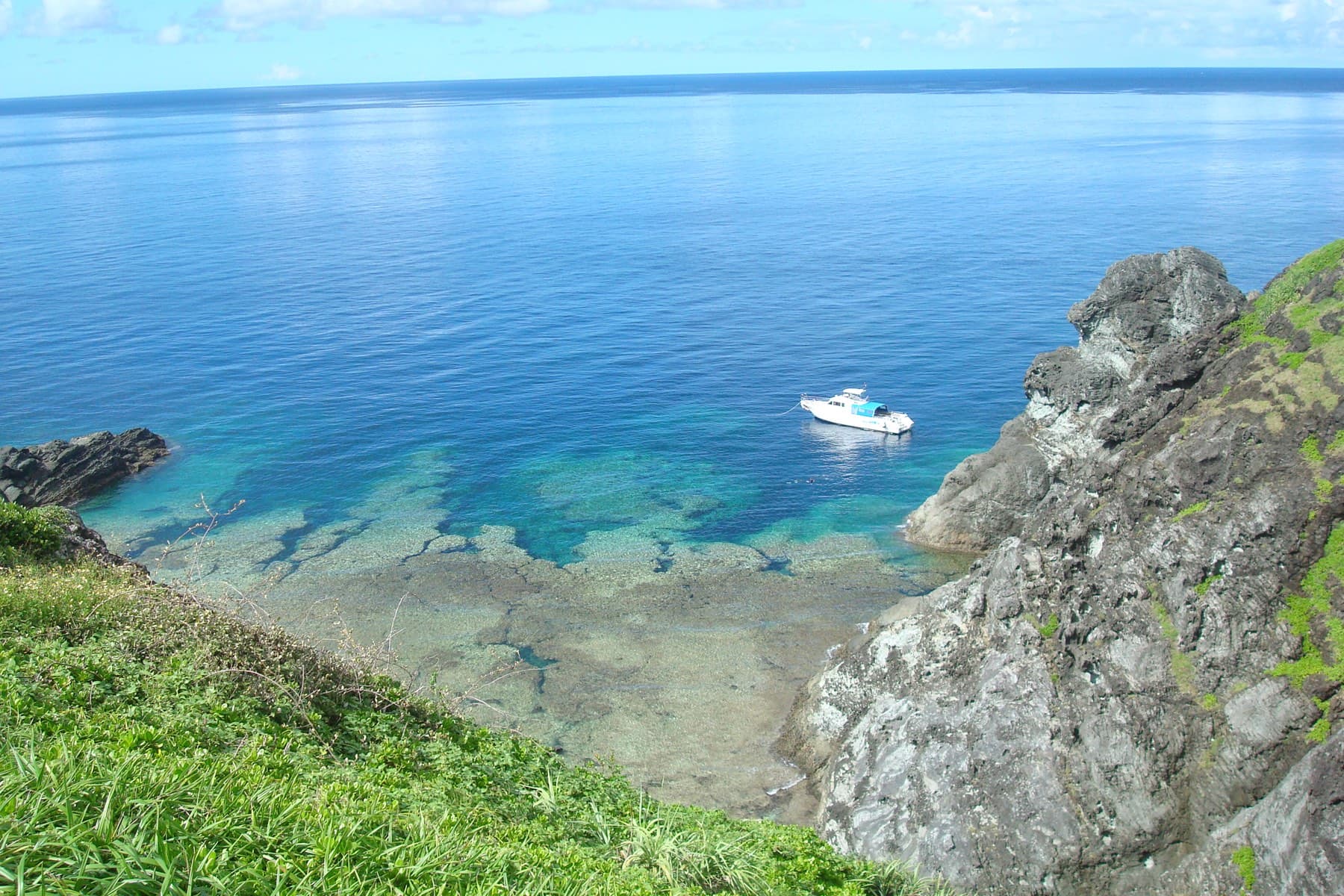 A small boat floats over clear reef water in a cove below the dark volcanic cliffs at Ogan-zaki on the west coast of Ishigaki.