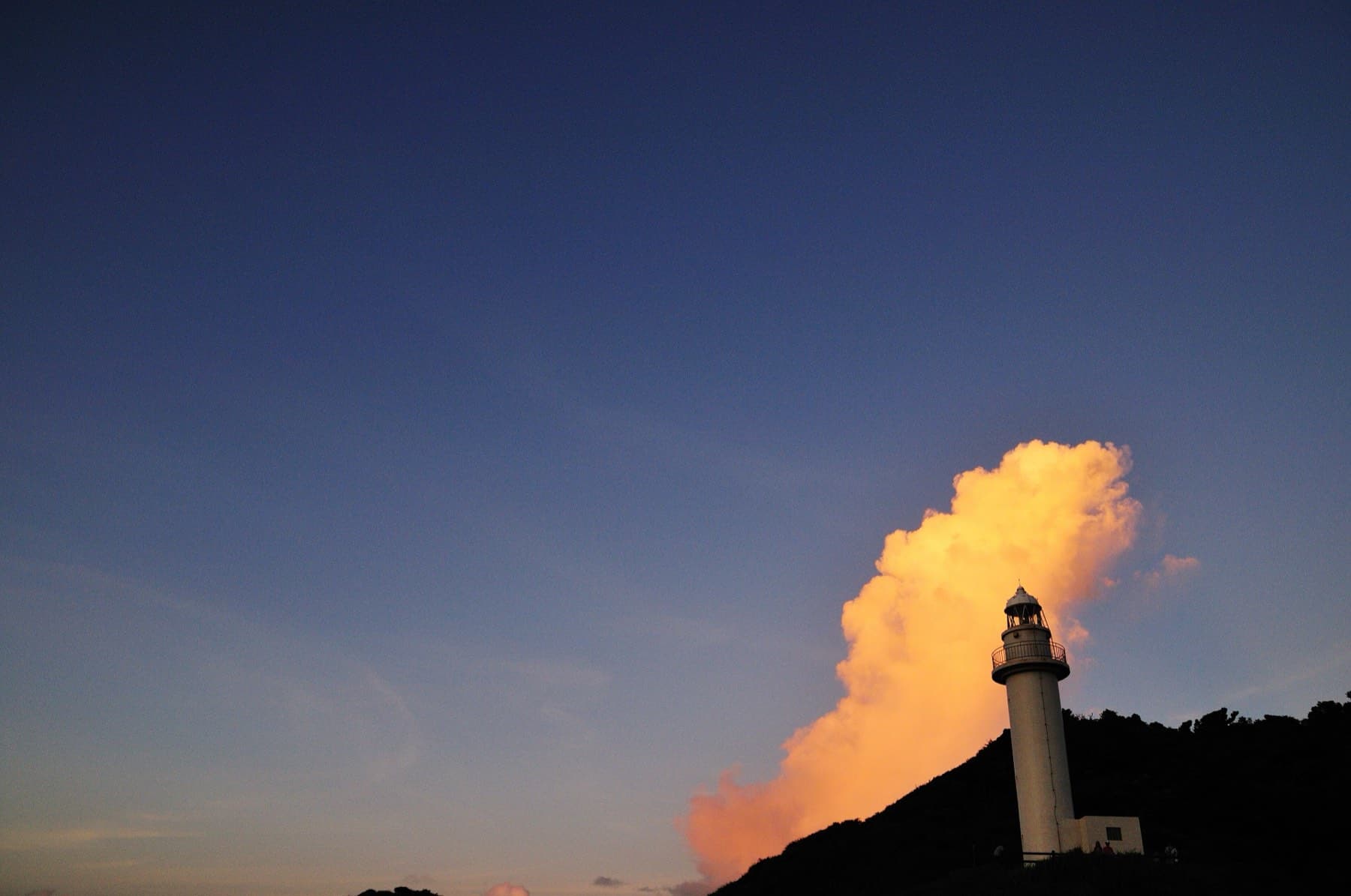 At dusk the Ogan-zaki lighthouse stands dark on its ridge while a tower of cloud behind it glows orange in the last of the sun.