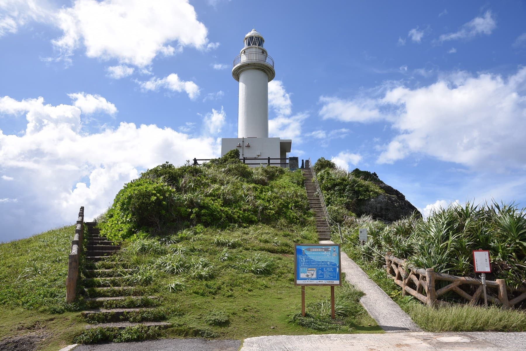 Steps and a path climb a grassy knoll to the white Ogan-zaki lighthouse at the western end of Ishigaki under a bright summer sky.