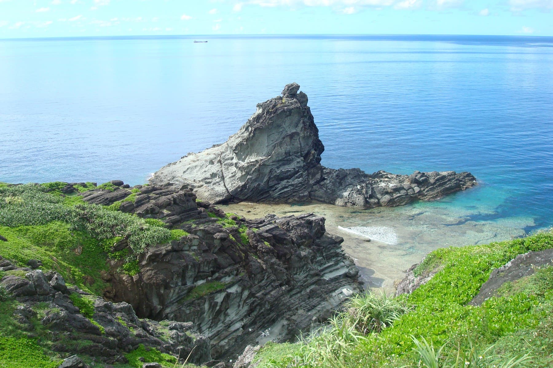 A jagged rock spire rises from the tilted strata of the Ogan-zaki headland above a pocket of sand and calm open sea.
