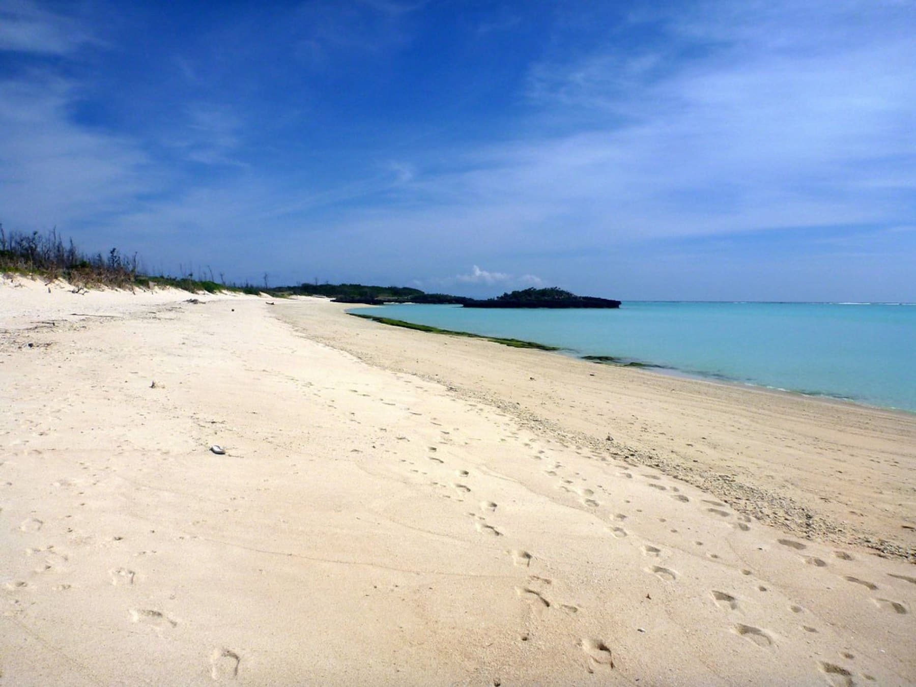 Footprints cross the long white sand of Oganeku Beach as the shallow water runs out toward a low rocky islet.
